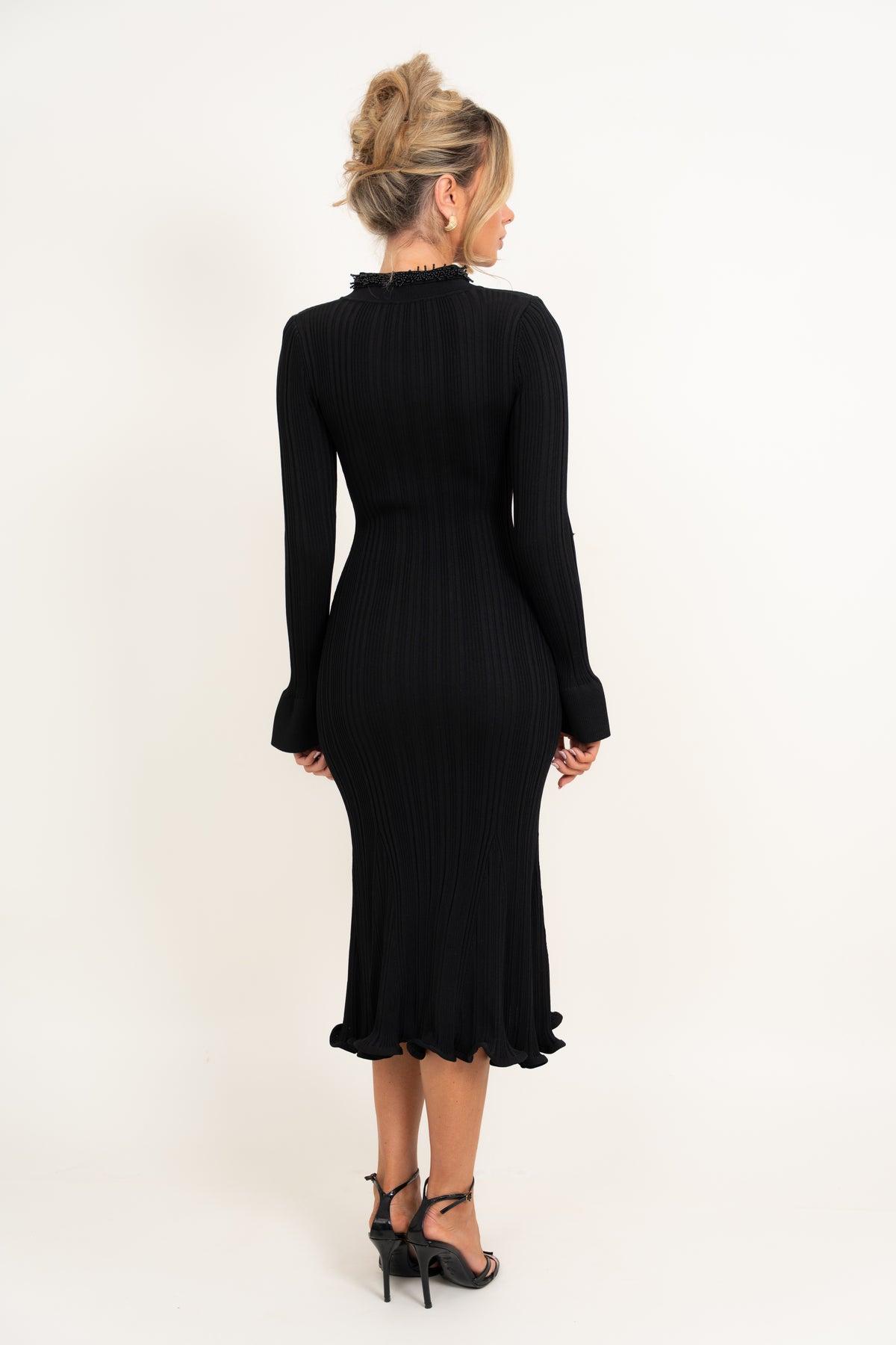 A woman viewed from the back wearing a black long-sleeve ribbed midi dress with a ruffled high neckline and ruffled hem, paired with black high-heeled sandals, against a plain white studio background.