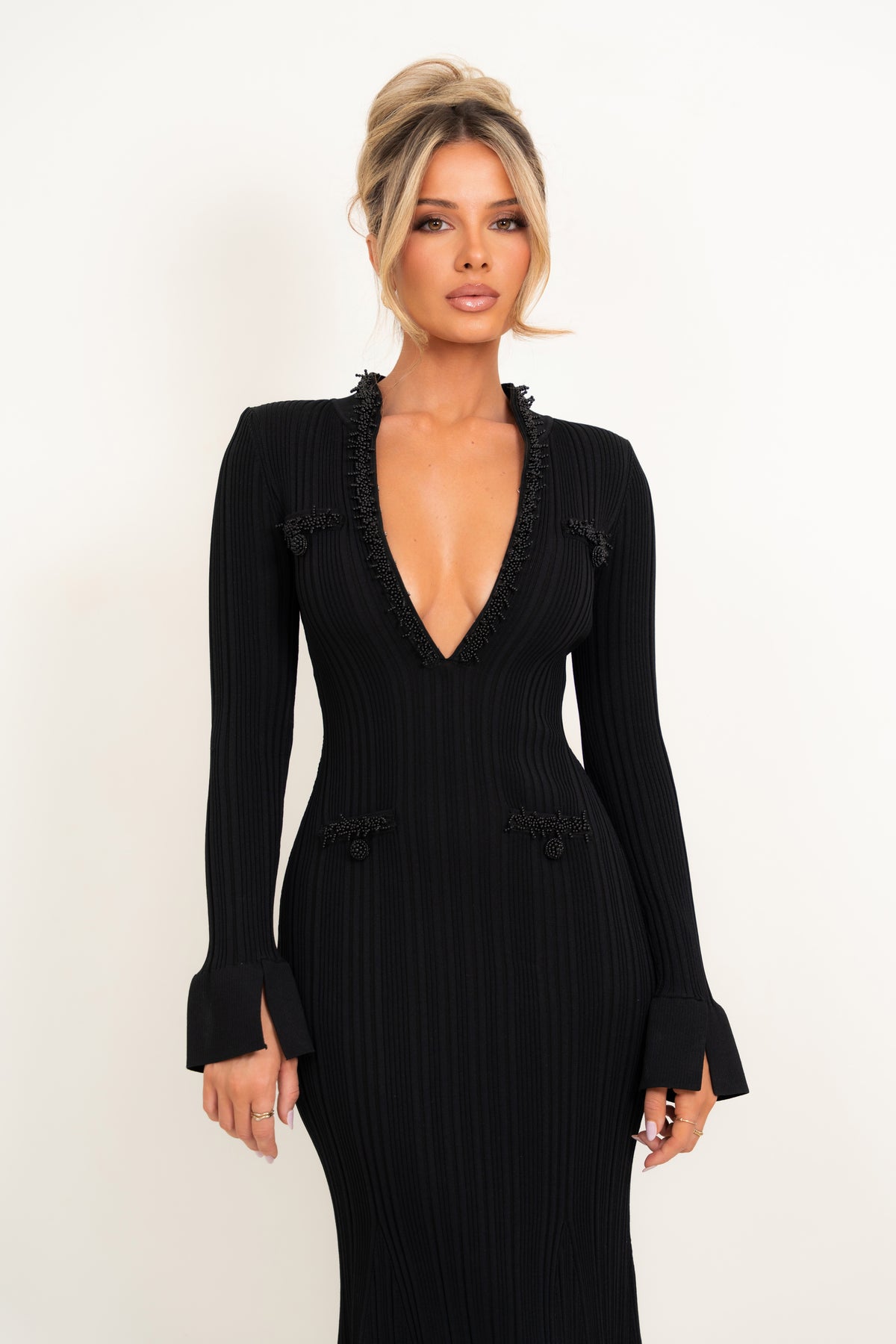 A woman modeling a black ribbed long-sleeve dress with a deep V-neckline, ruffled trim, and decorative buttons on the shoulders and waist, set against a plain white studio background.