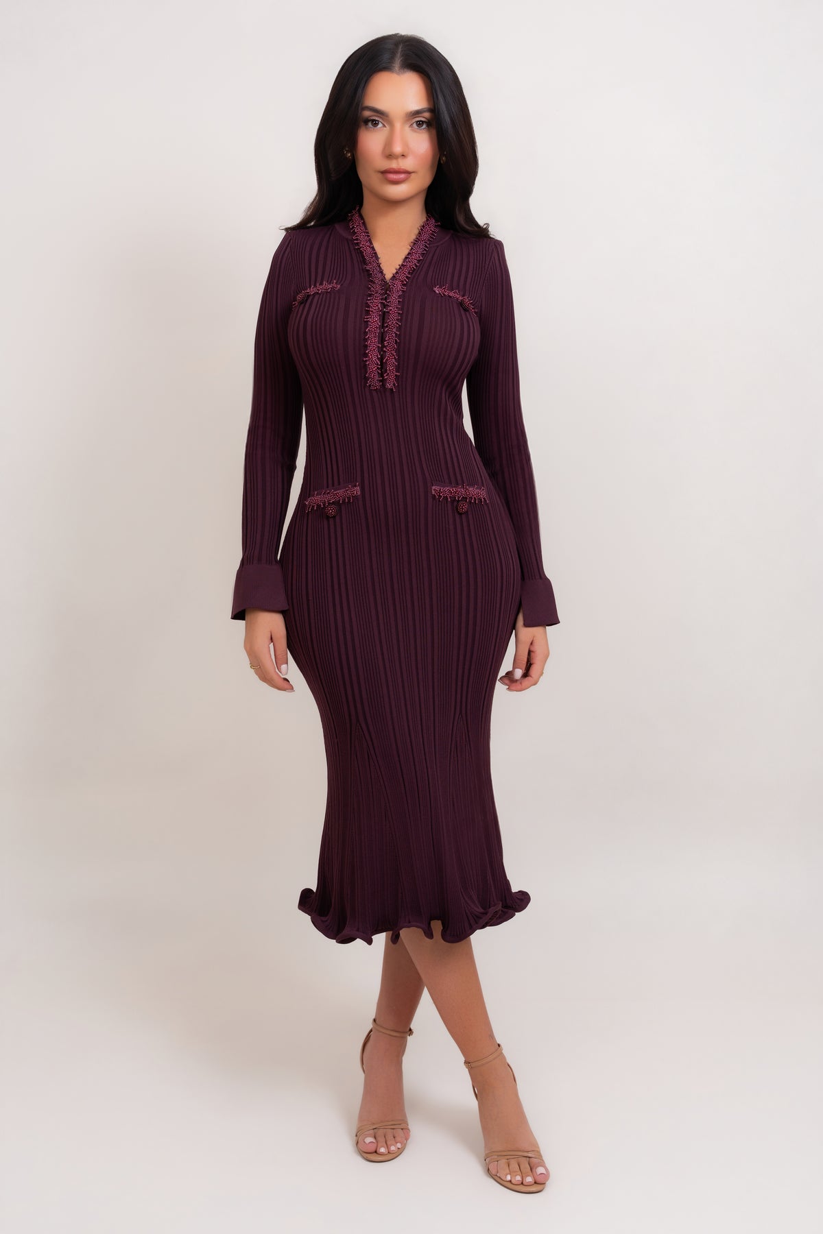 A woman modeling a long-sleeve, dark plum purple pleated midi dress with ruffle trim details on the neckline and pockets, paired with beige high-heeled sandals against a plain light background.
