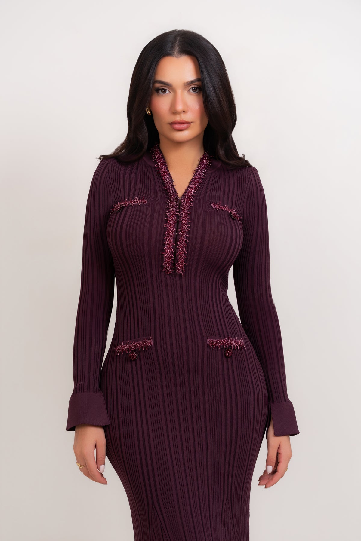 A woman modeling a long-sleeve ribbed knit dress in deep plum purple with matching fringed trim details on the neckline and pockets, set against a plain light gray studio background.