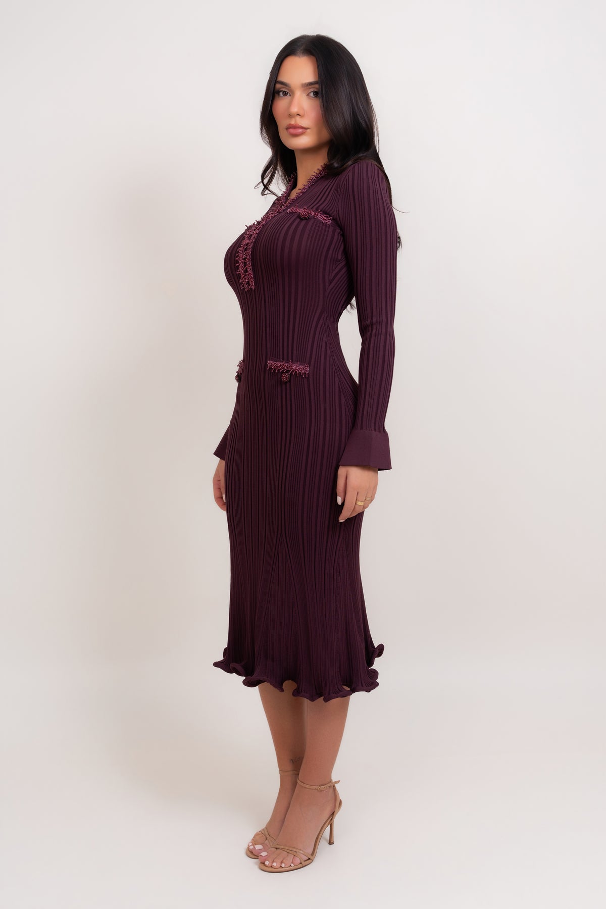 A woman modeling a long-sleeve, dark plum purple ribbed knit midi dress with ruffle trim details, standing against a plain off-white studio background.