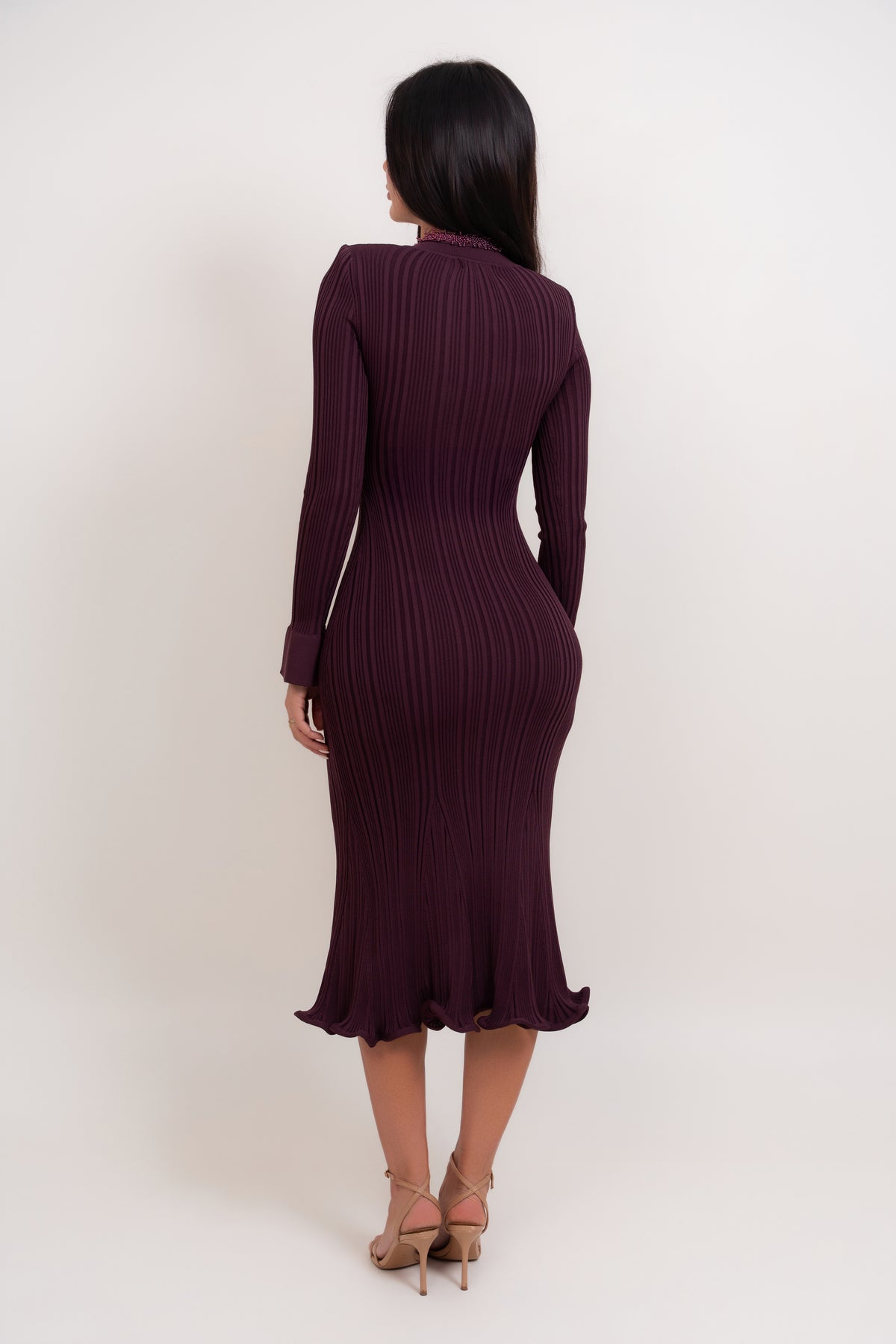 A woman viewed from the back wearing a dark plum ribbed long-sleeve midi dress with a ruffled hem, paired with beige high-heeled sandals, against a plain off-white studio background.