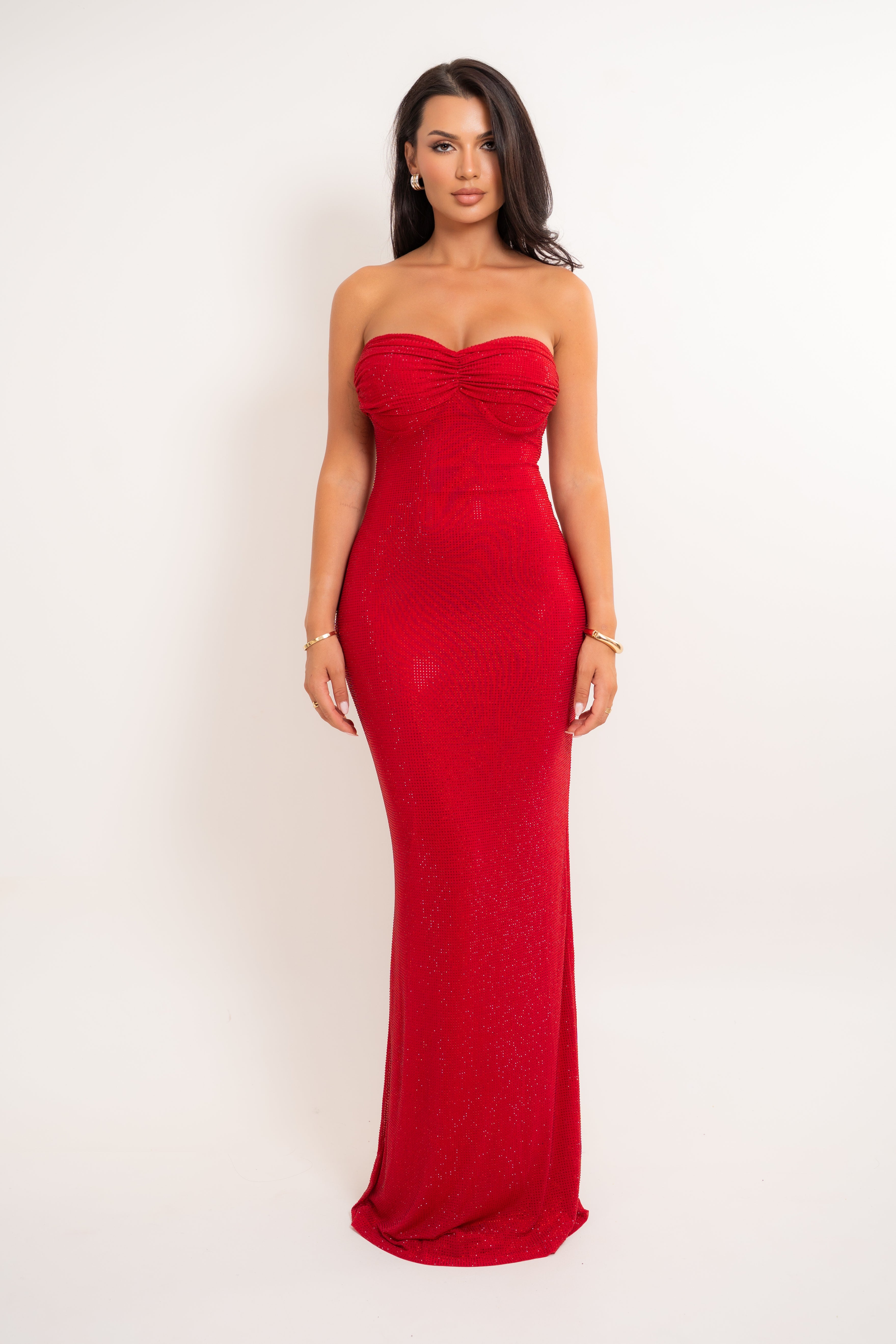 A woman modeling a strapless red maxi gown with a ruched sweetheart neckline and a fitted silhouette, photographed against a plain light background.