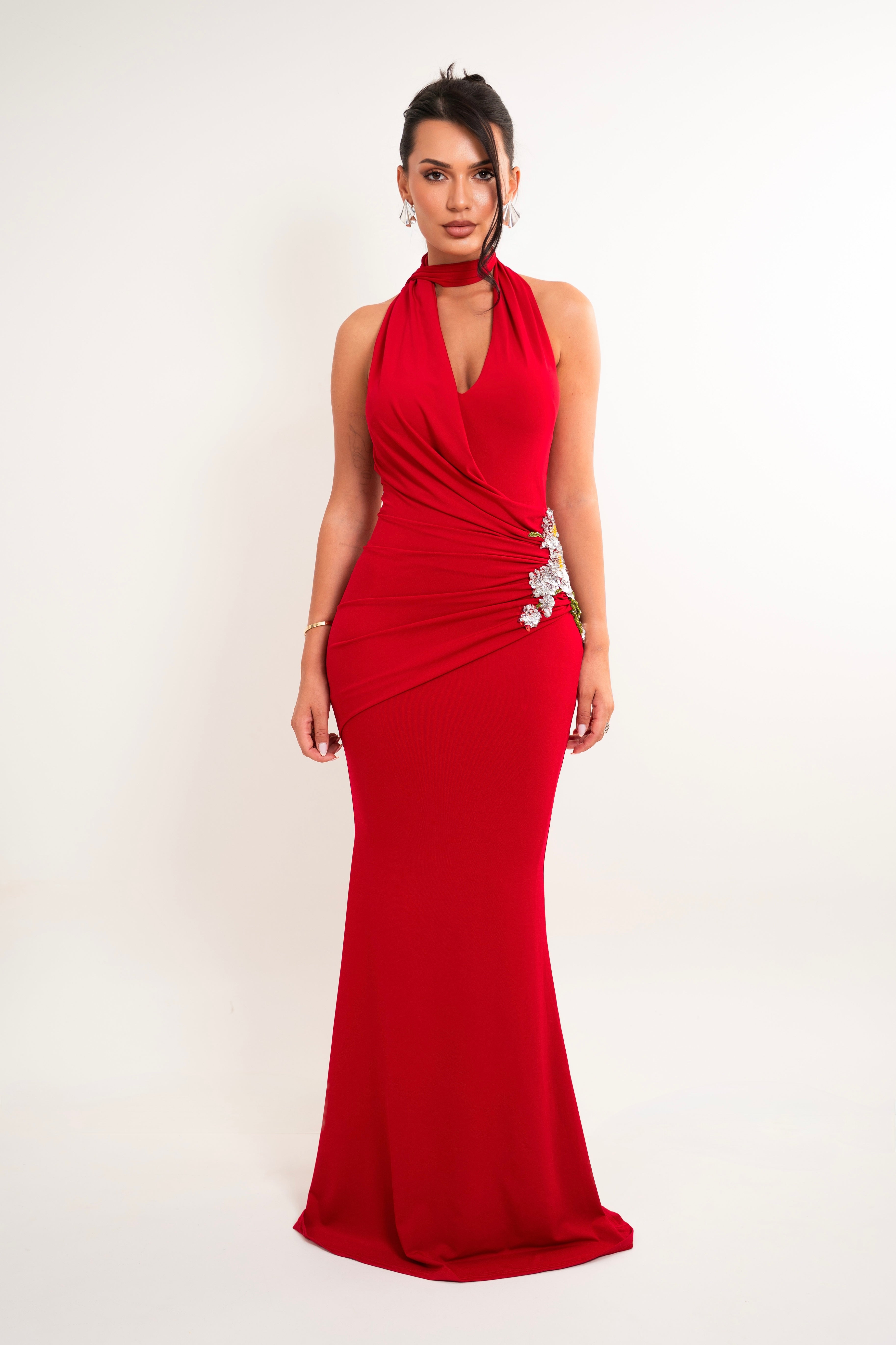 A woman modeling a floor-length red halter gown with a draped wrap-style bodice and a jeweled embellishment at the waist, photographed against a plain light background.