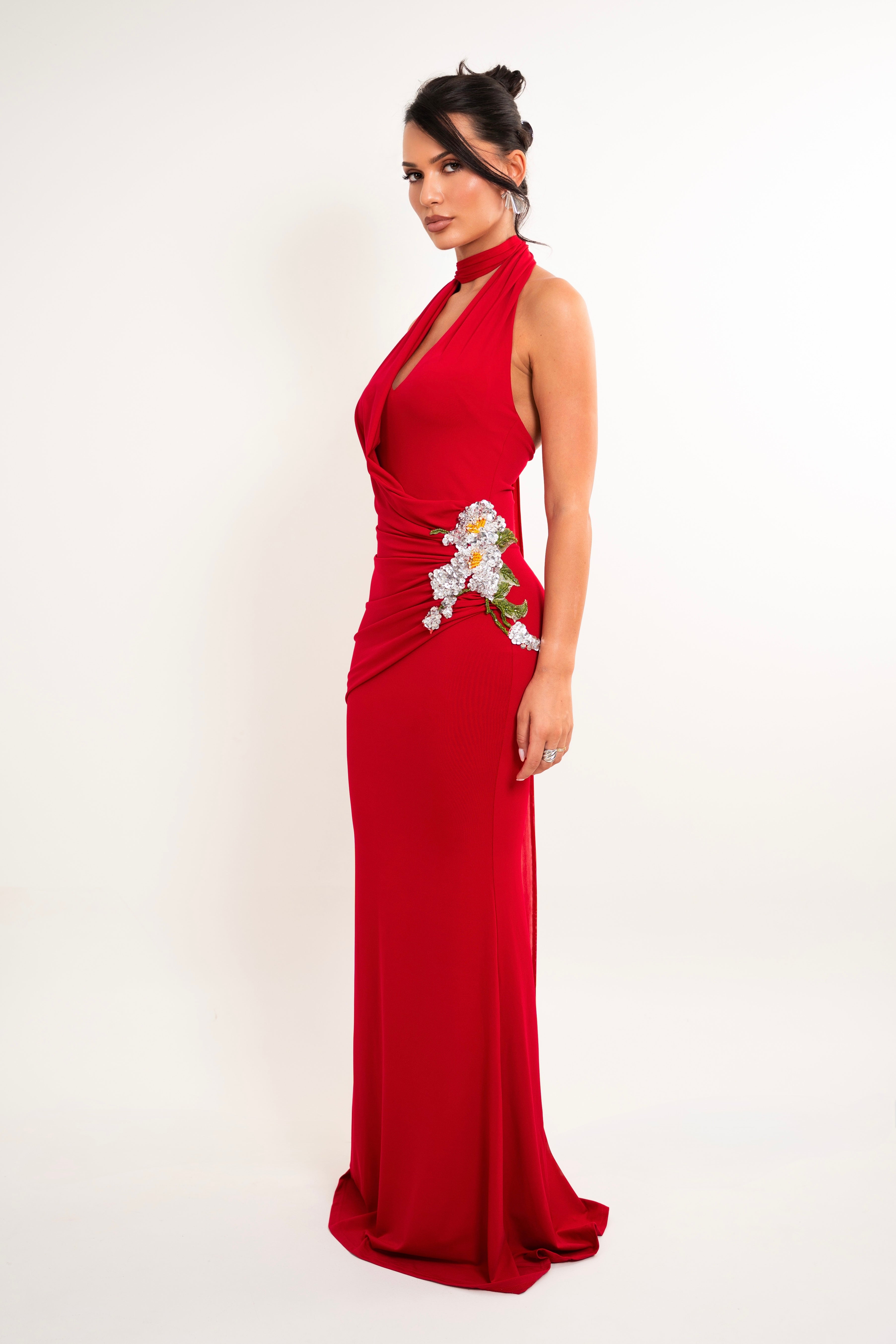A woman modeling a floor-length red halter gown with a draped wrap-style bodice and a floral applique detail at the waist, photographed against a plain white studio background.