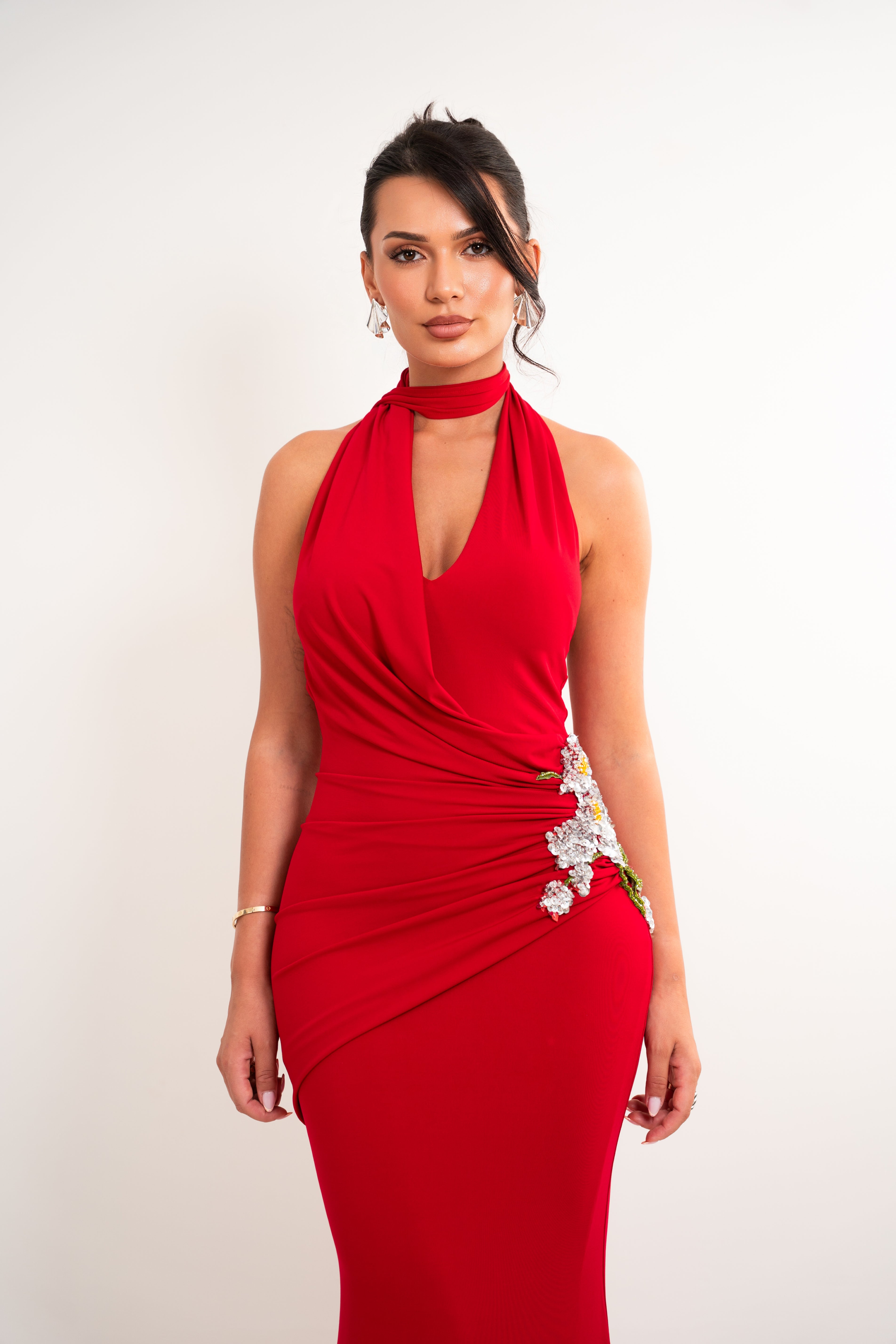 A woman modeling a bright red halter-neck evening gown with a keyhole cutout at the chest, ruched draping details, and white floral embellishments on the side, set against a plain light background.
