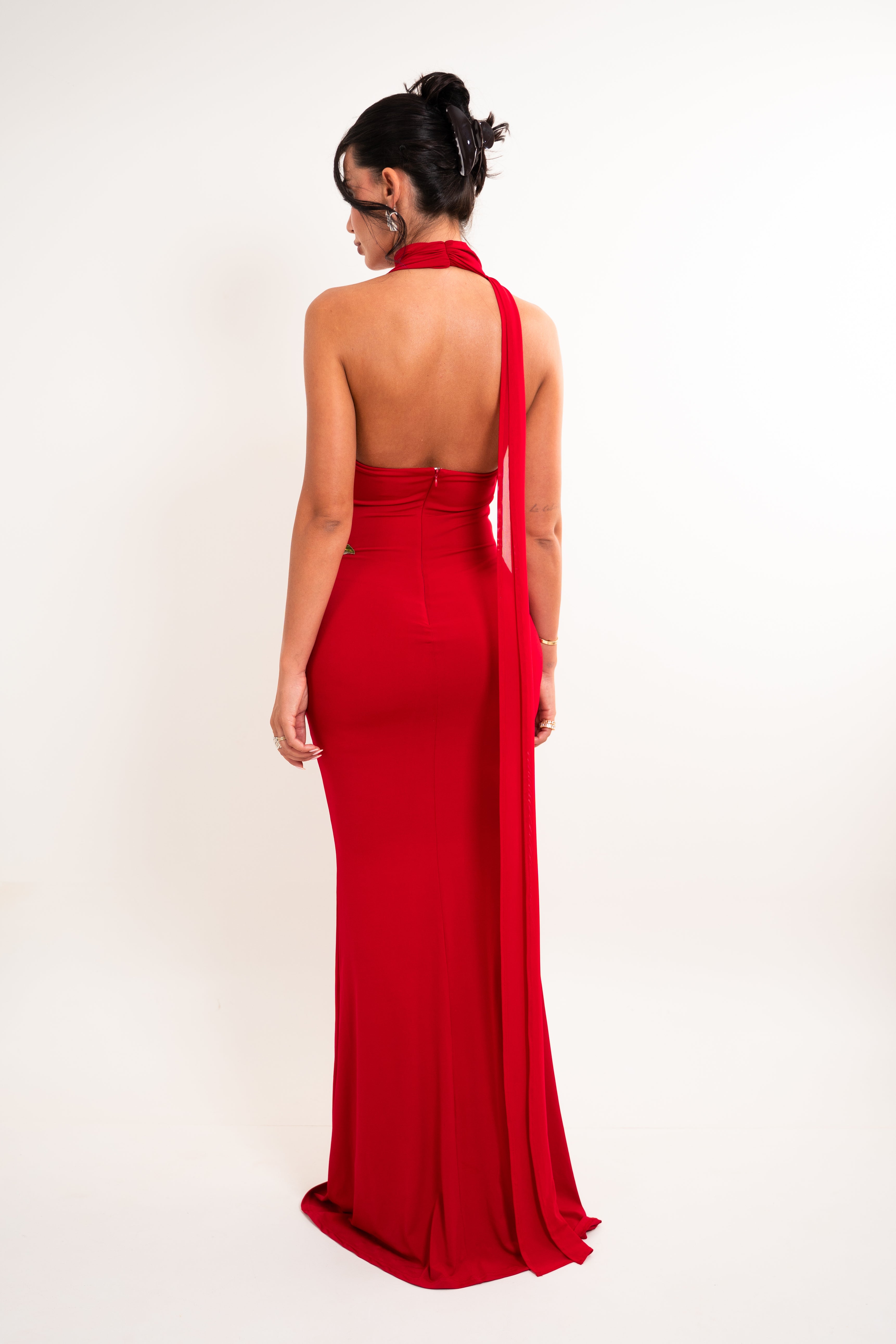 A woman viewed from the back wearing a bright red halter-neck evening gown with a long draped train against a plain light background.