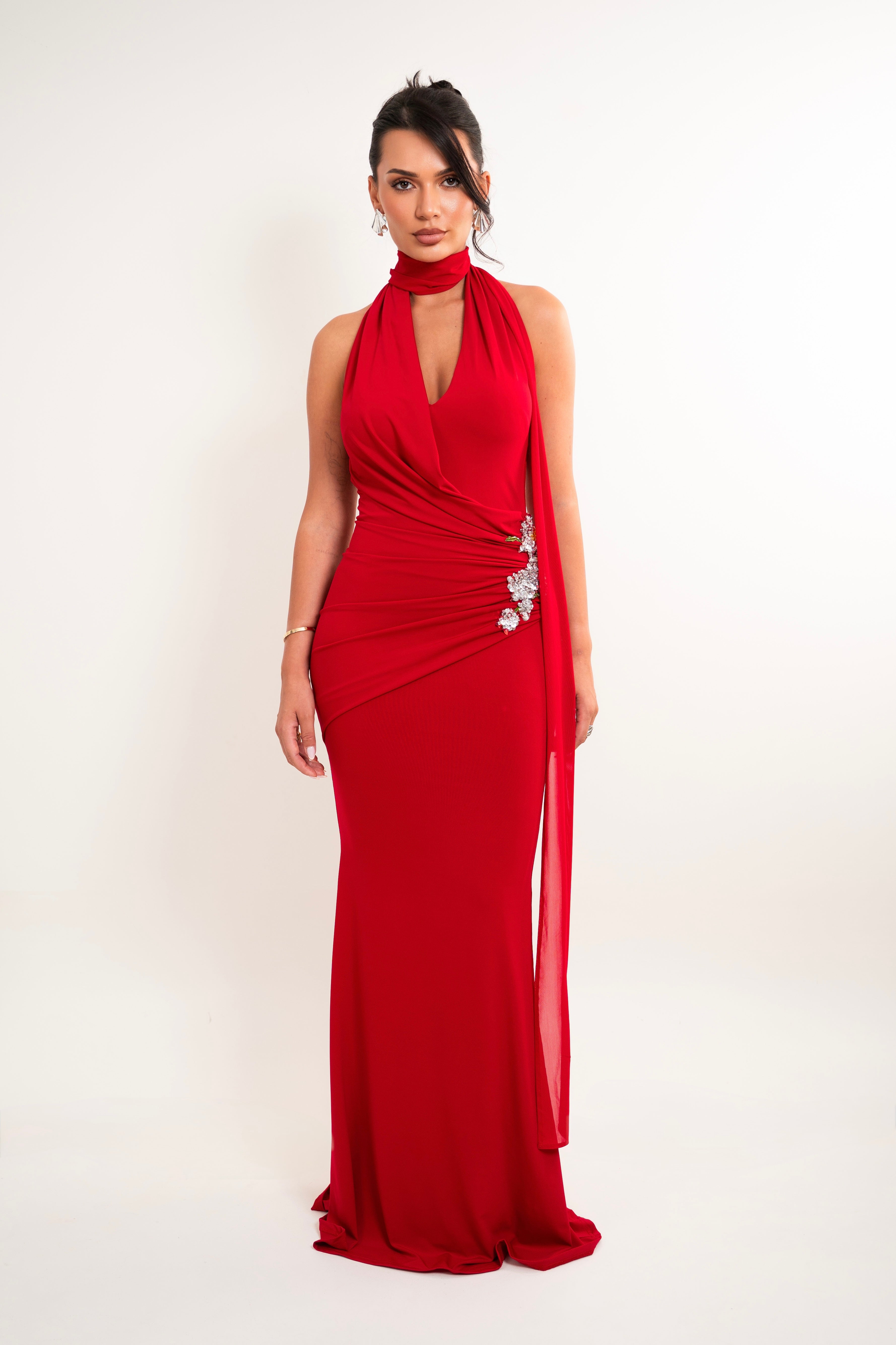 A woman modeling a floor-length red evening gown with a halter neckline, draped side detail, and a jeweled embellishment at the waist, photographed against a plain light background.