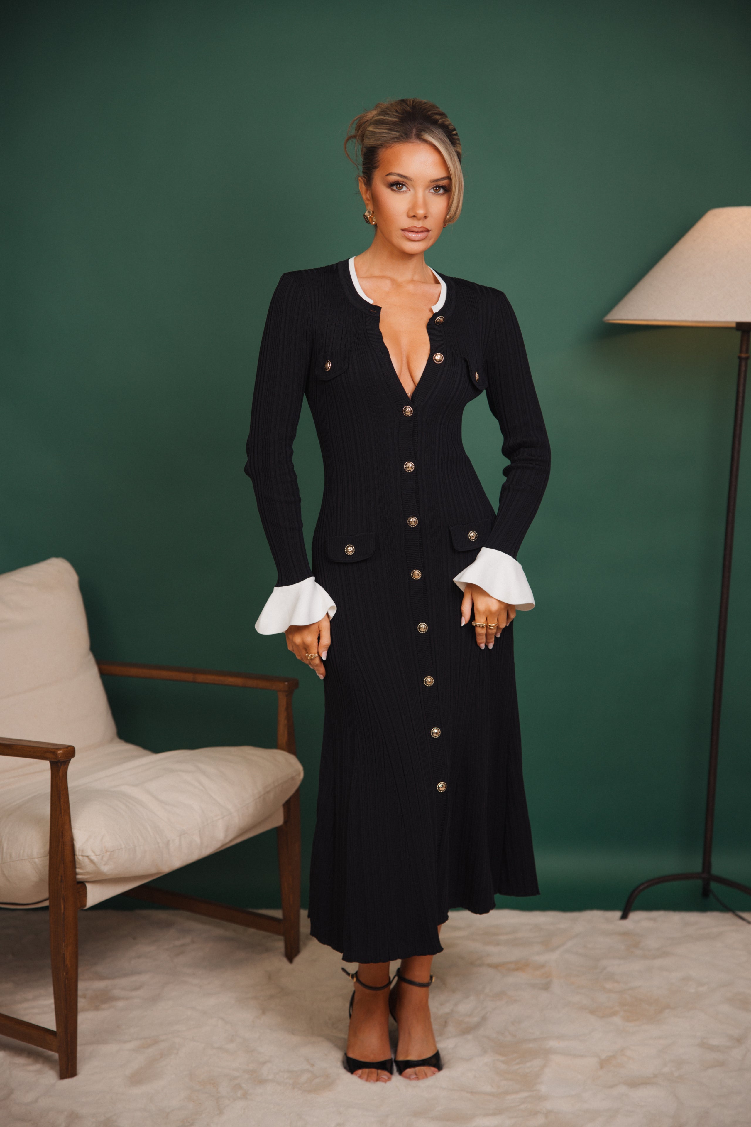 Woman in a black dress with white cuffs standing against a green wall.