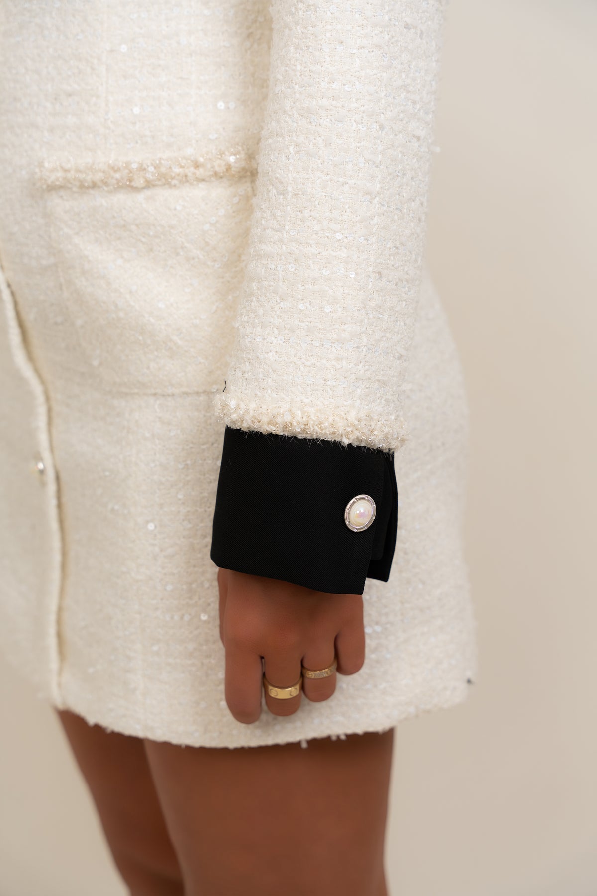 Close-up of a person wearing a cream tweed jacket with a contrasting black cuff and a single white pearl button, with gold rings on the hand.