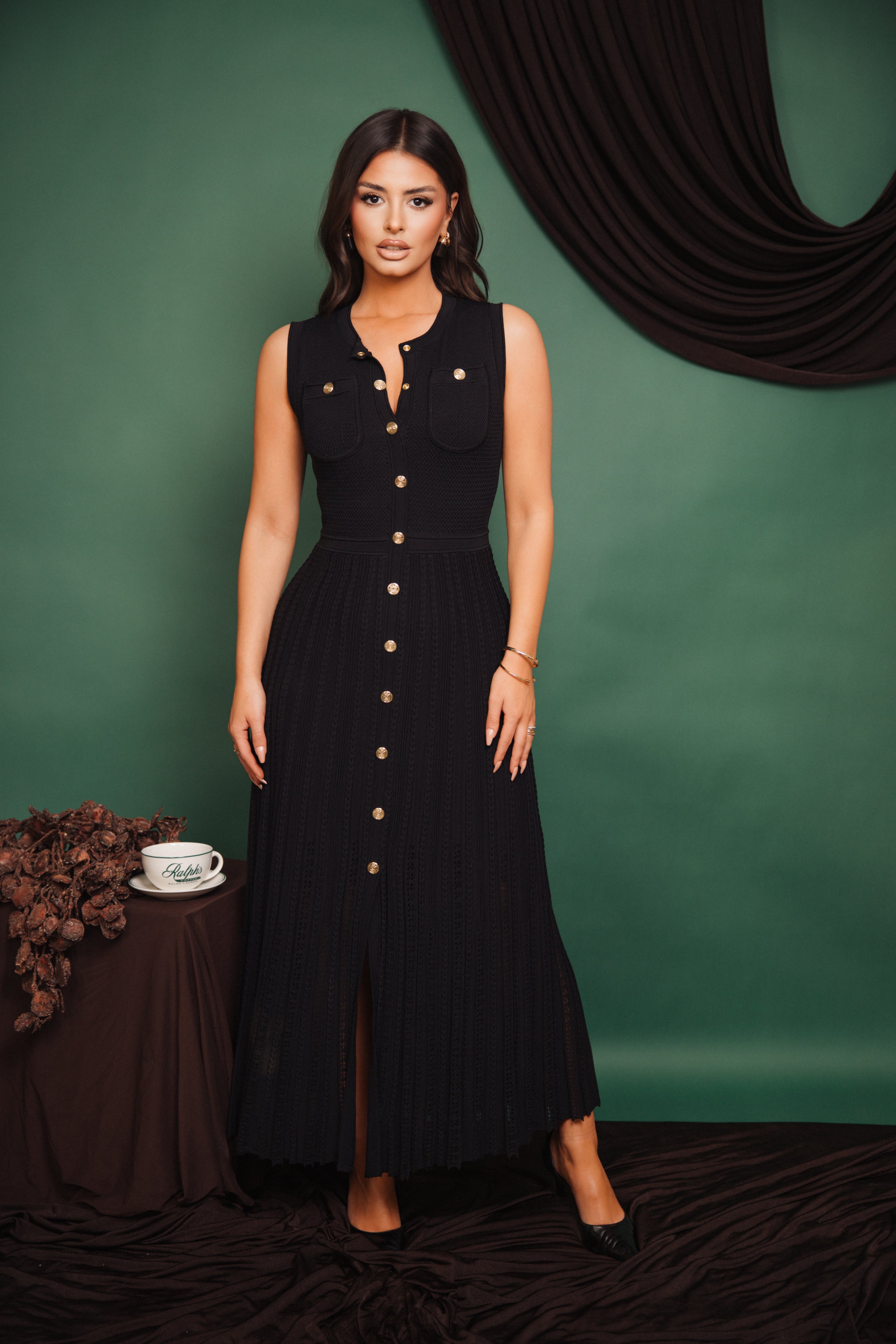 Woman in a black dress standing against a green background with a table and decorative elements.
