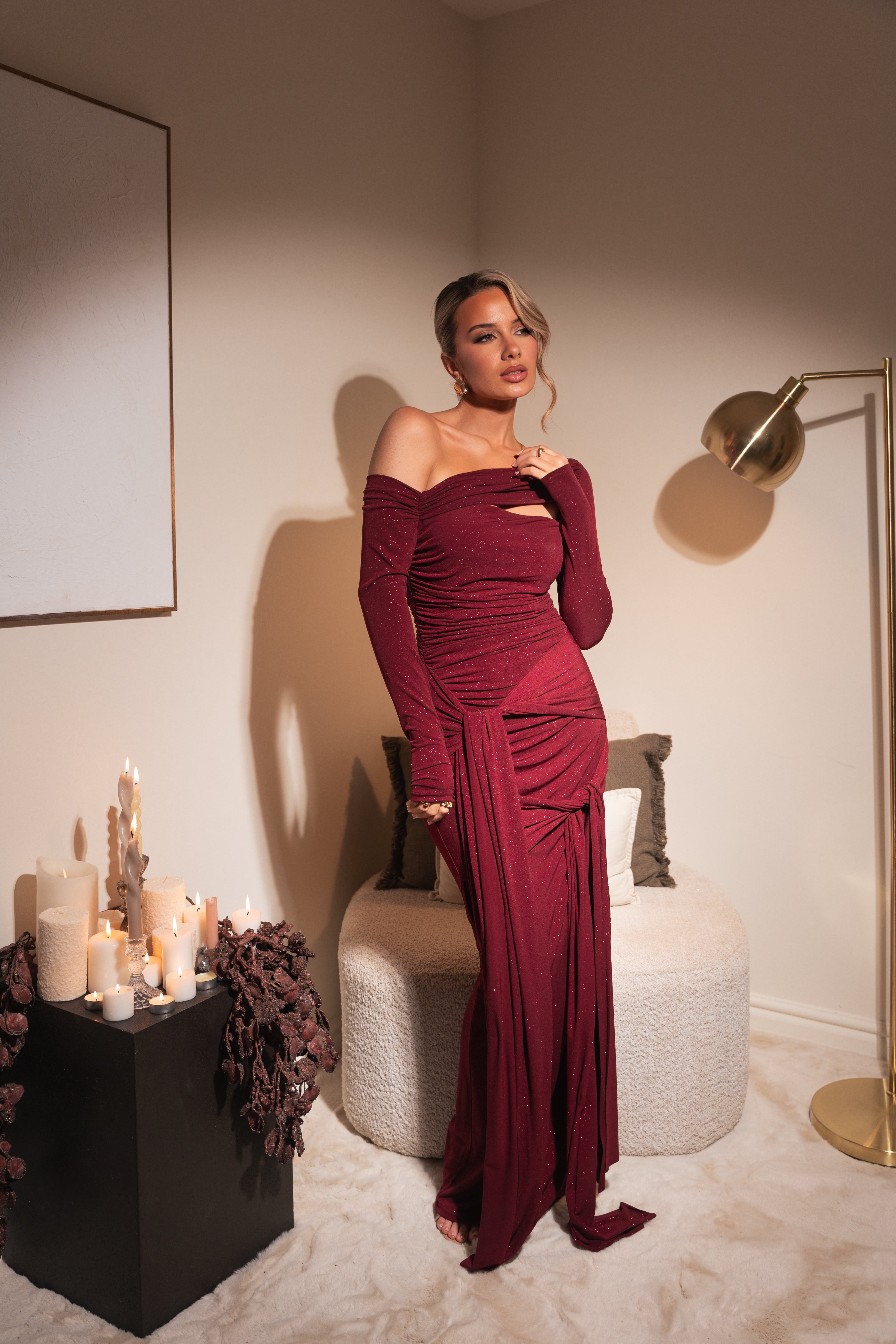 Woman in a burgundy off-shoulder gown standing in a room with candles and decor.