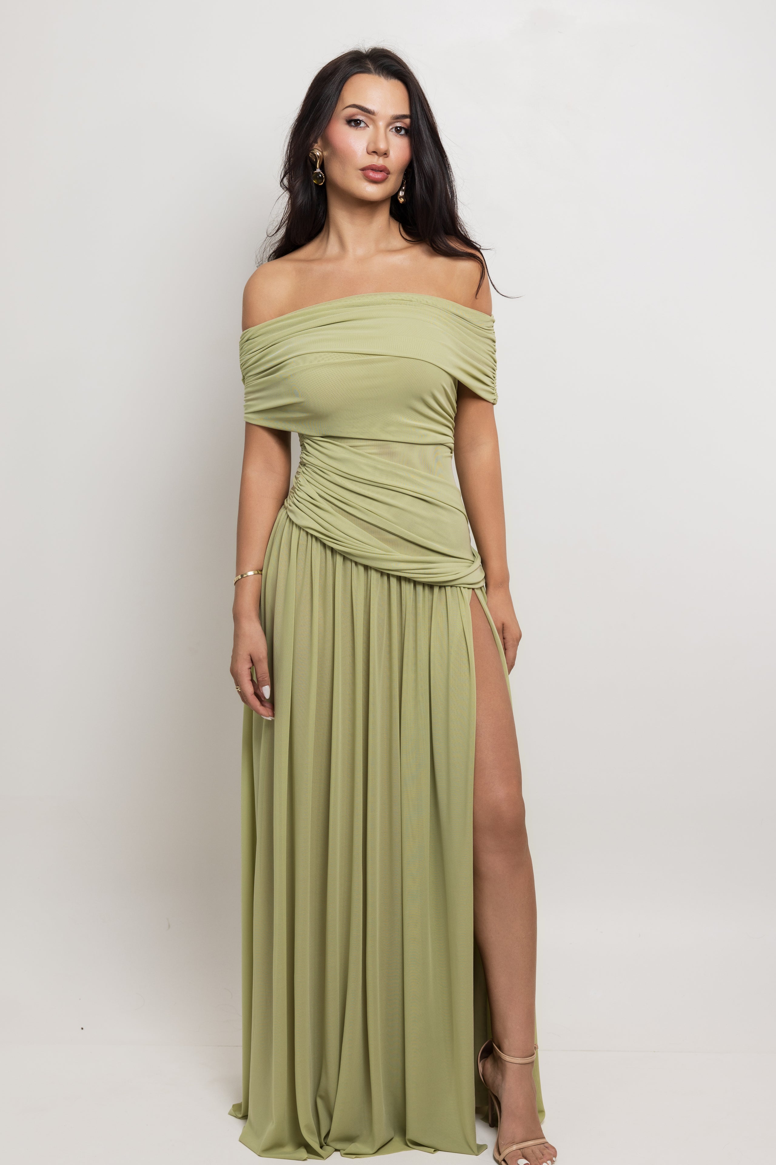A woman modeling an off-the-shoulder olive green maxi dress with a ruched bodice, draped sleeves, and a high thigh slit, set against a plain light gray studio background.