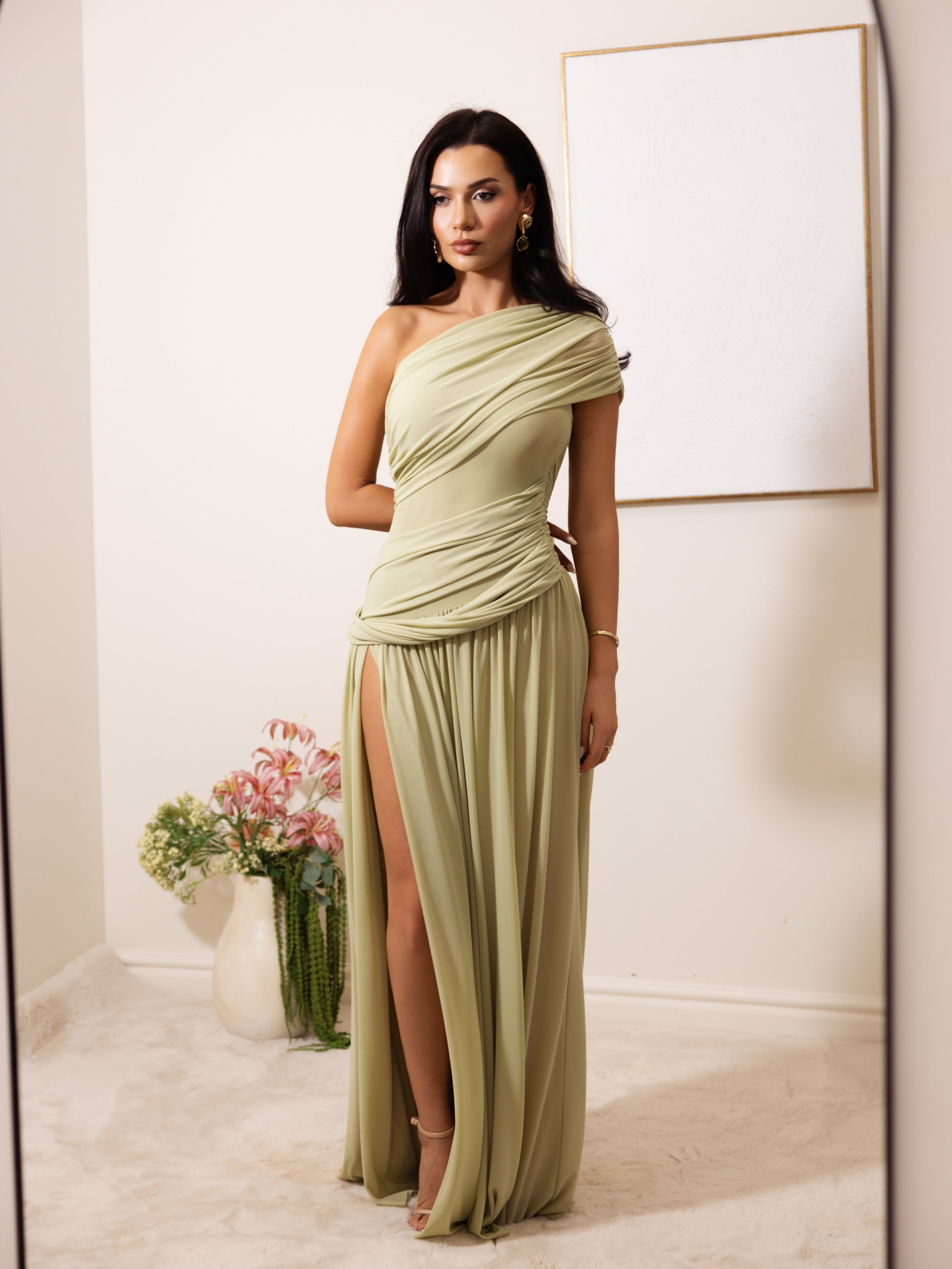 A woman models a floor-length draped gown in a soft sage green color, featuring an asymmetrical one-shoulder design, a high thigh slit, and a gathered waist. She is standing in a minimalist room with a white wall, a blank framed picture, and a vase of flowers.