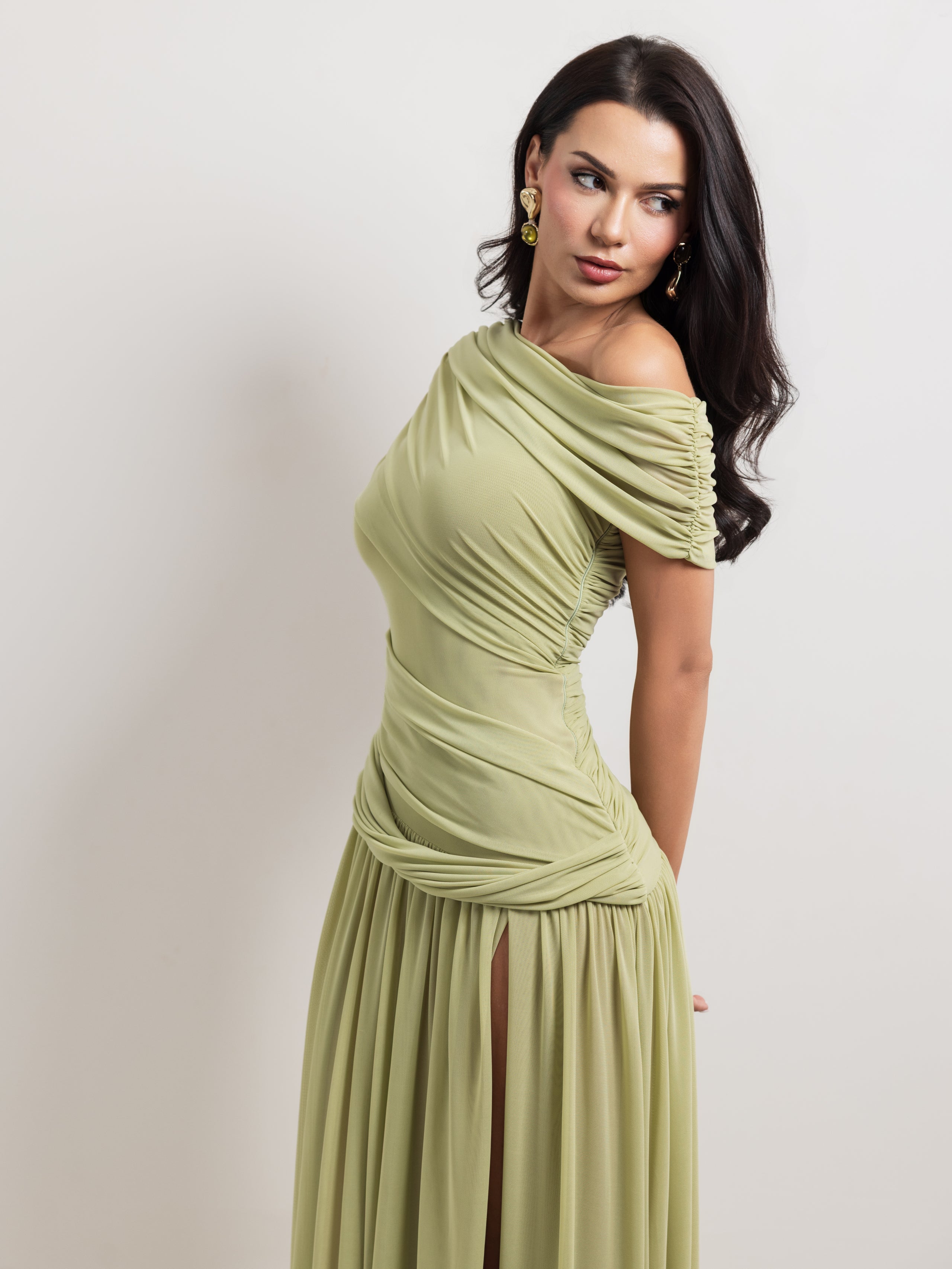 A woman modeling an off-the-shoulder, draped maxi dress in a light sage green color, posed against a neutral light gray studio background.