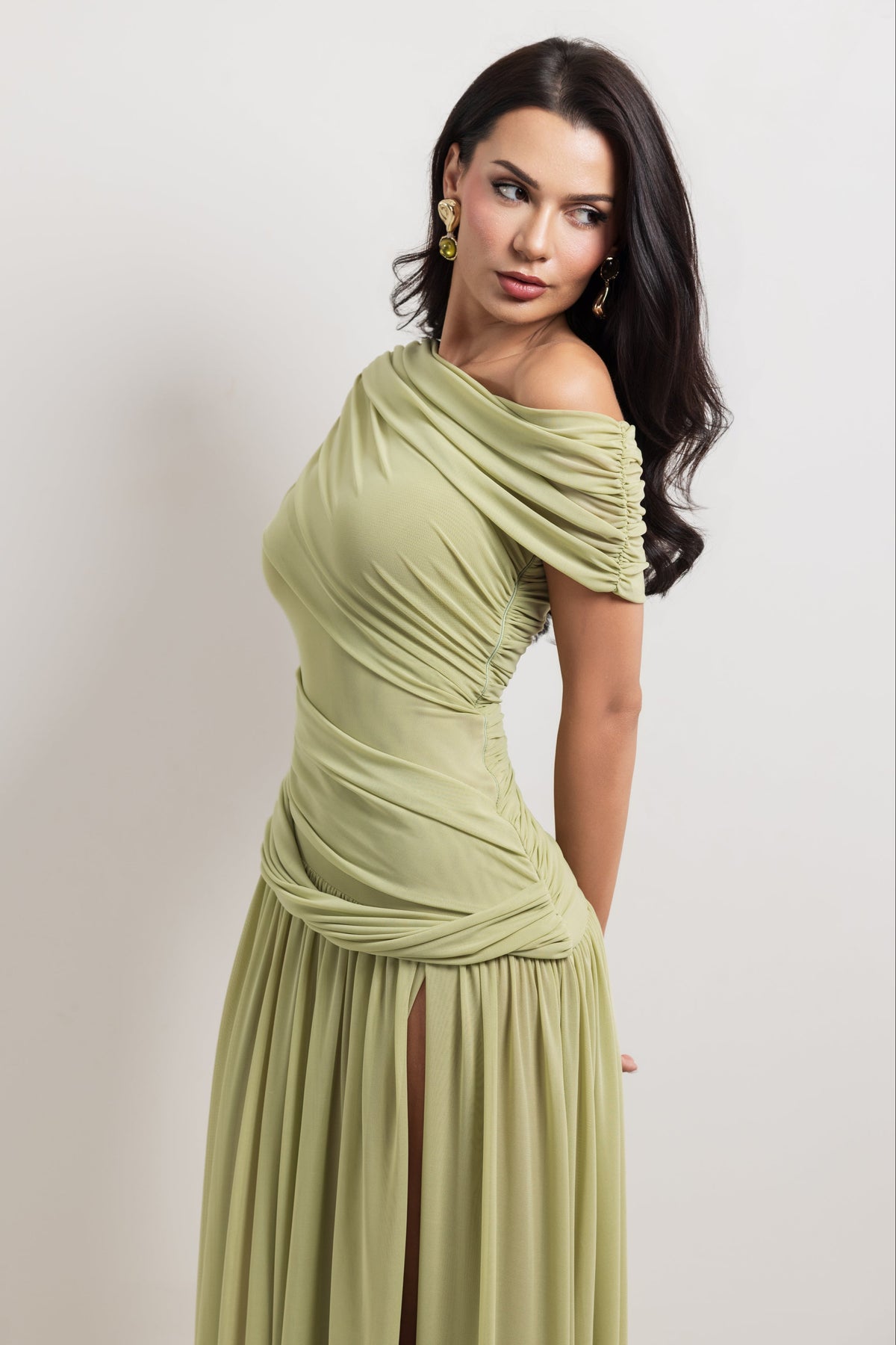 A woman modeling an off-the-shoulder, draped maxi dress in a light sage green color, posed against a neutral light gray studio background.