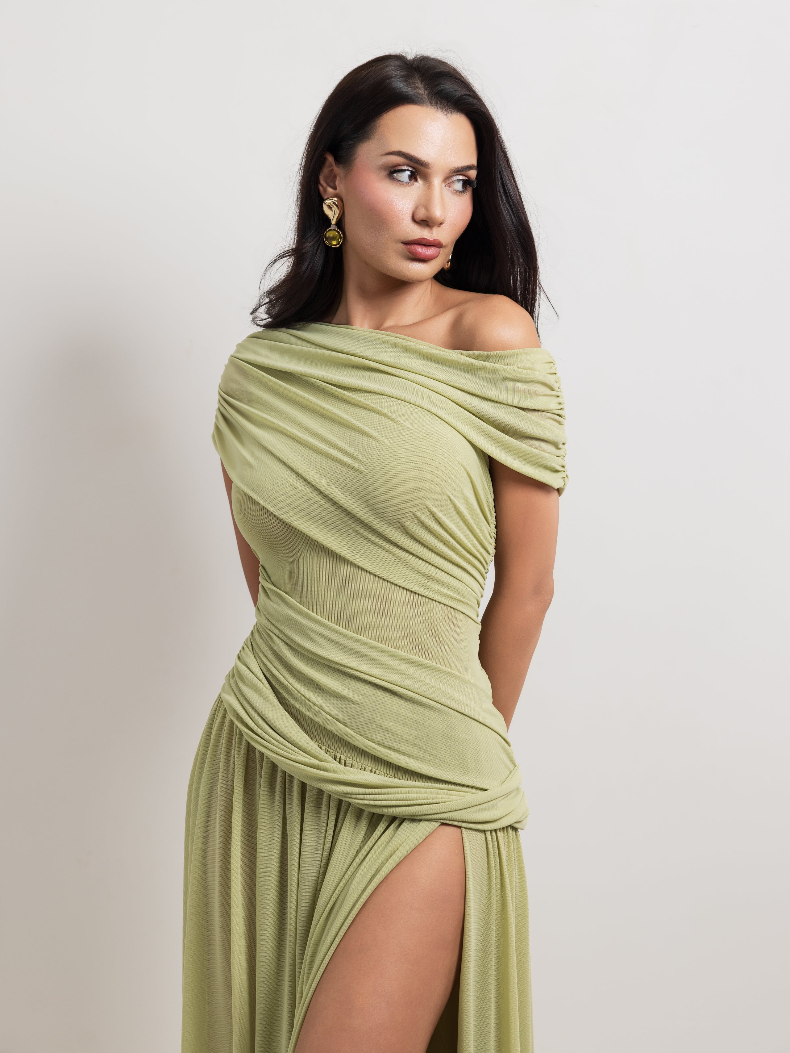 A woman modeling an off-the-shoulder sage green draped dress with a high thigh slit, photographed against a plain light gray studio background.