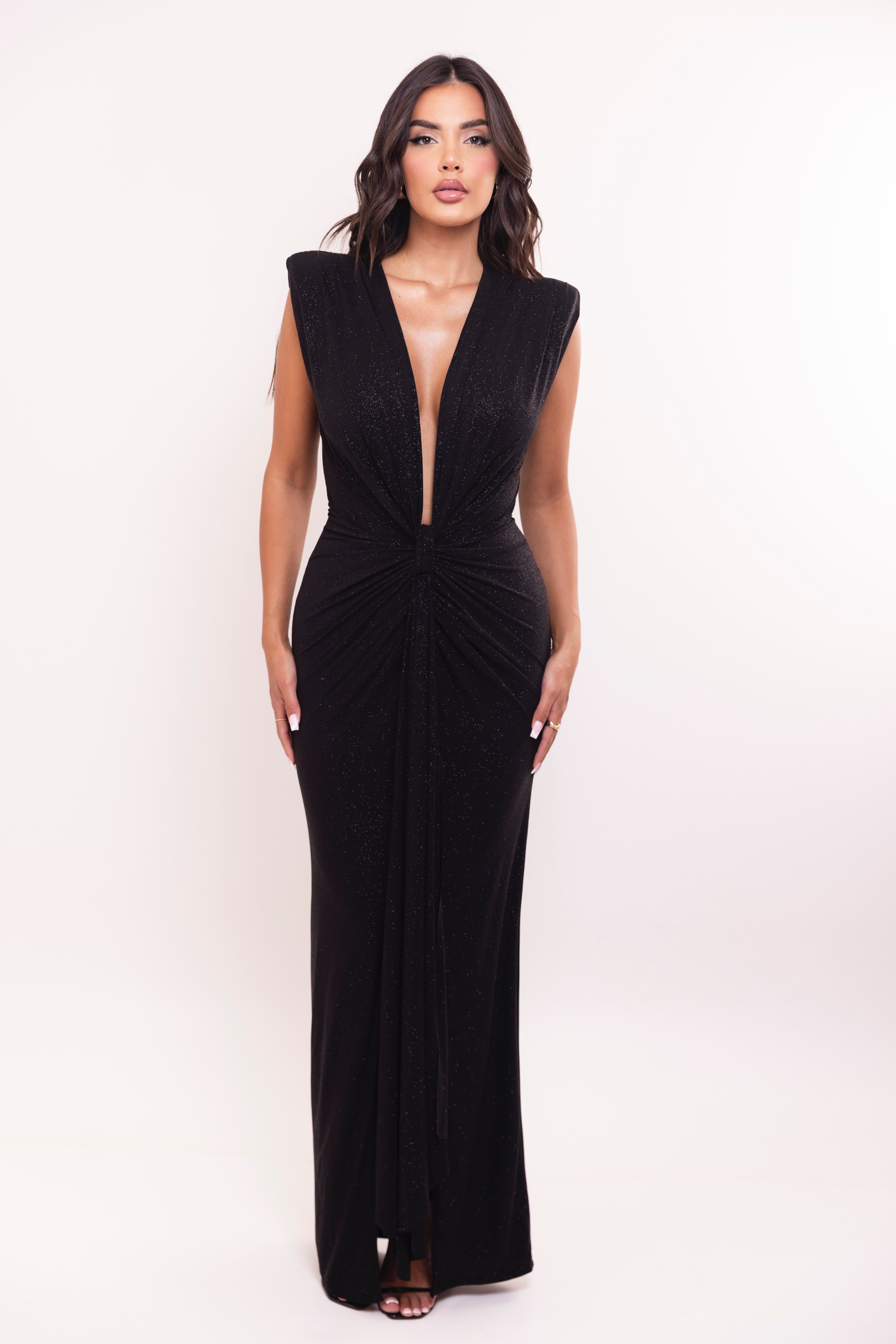 A woman modeling a sleeveless black maxi dress with a plunging V-neckline and gathered ruching at the waist, photographed against a plain light studio background.