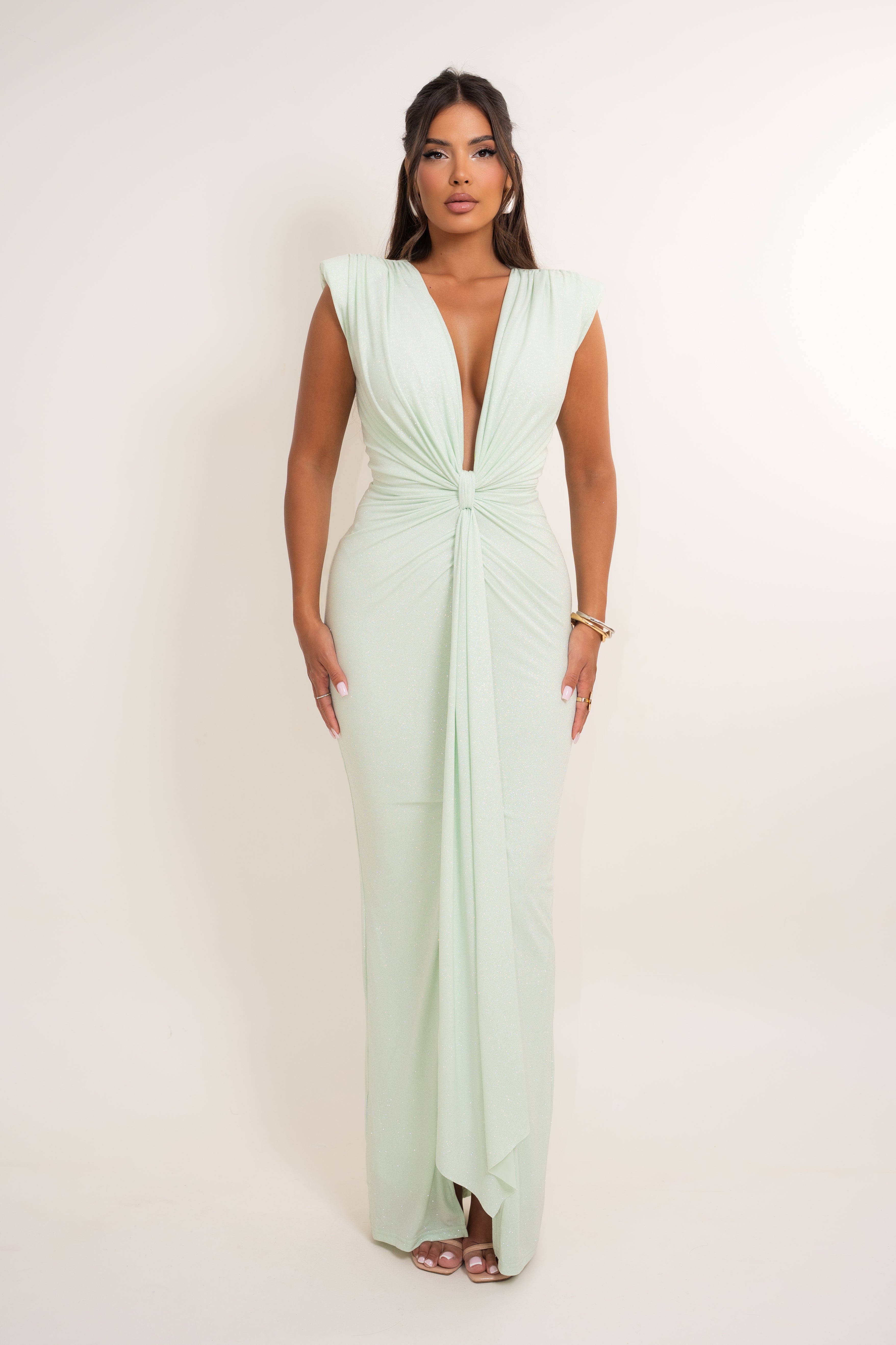 A woman modeling a floor-length sage green gown with a plunging V-neckline, cap sleeves, and a gathered twist detail at the waist, photographed against a plain off-white studio background.
