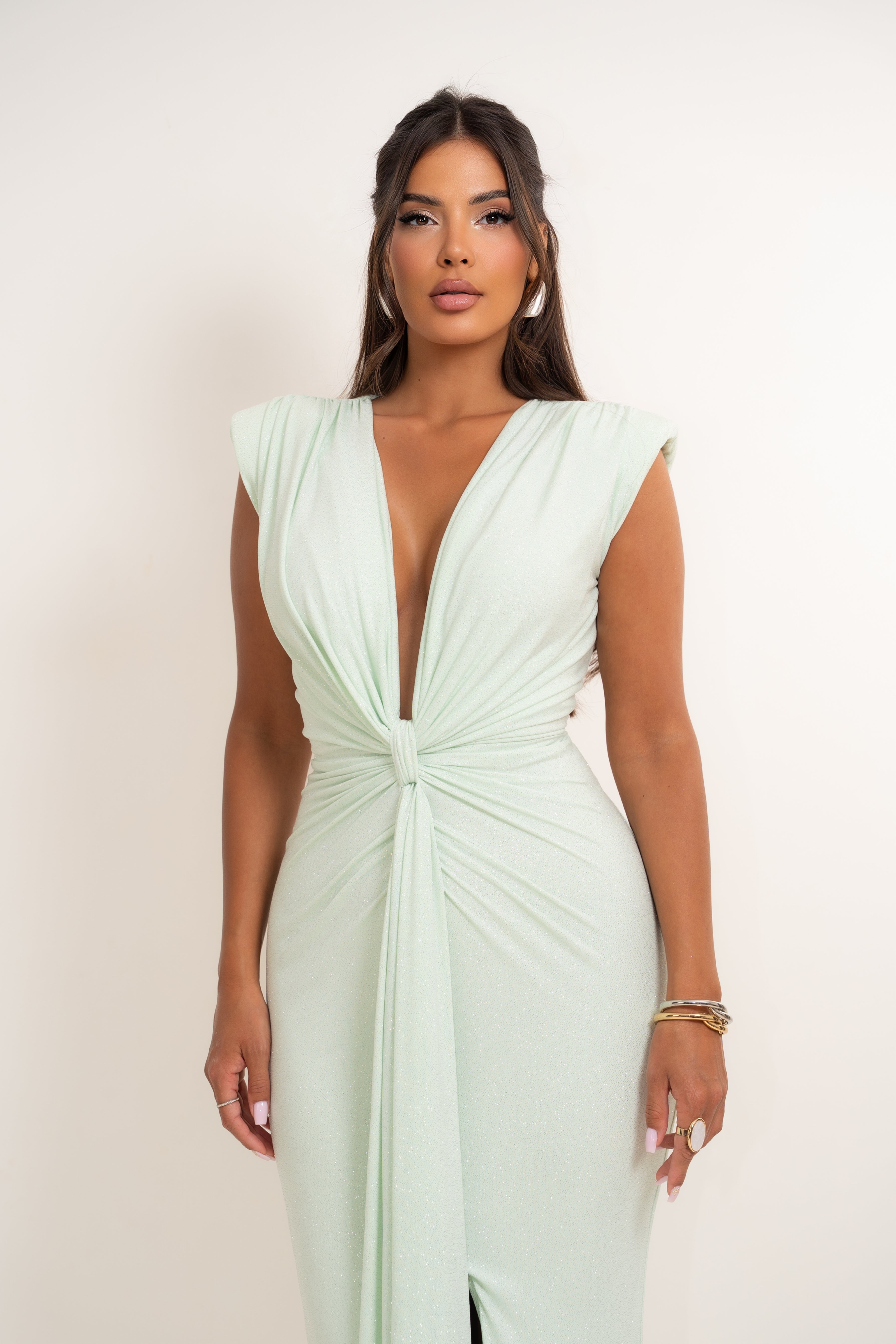 A woman modeling a pale mint green sleeveless maxi dress with a deep V-neckline and a twisted knot detail at the waist, set against a plain off-white studio background.