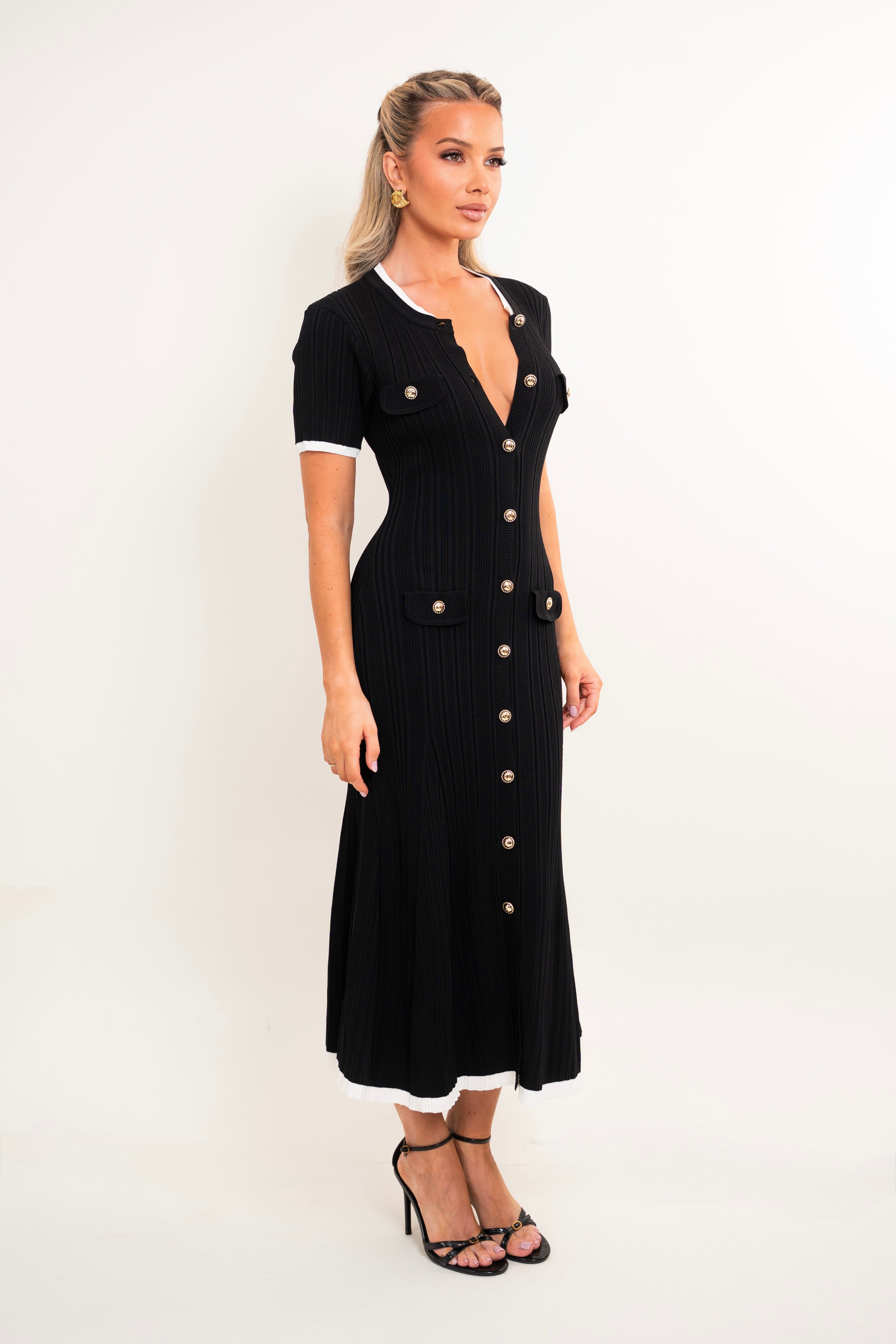 A woman modeling a black ribbed midi dress with white trim on the collar and sleeve cuffs, featuring gold-tone buttons down the front and on the chest pockets, set against a plain white studio background.
