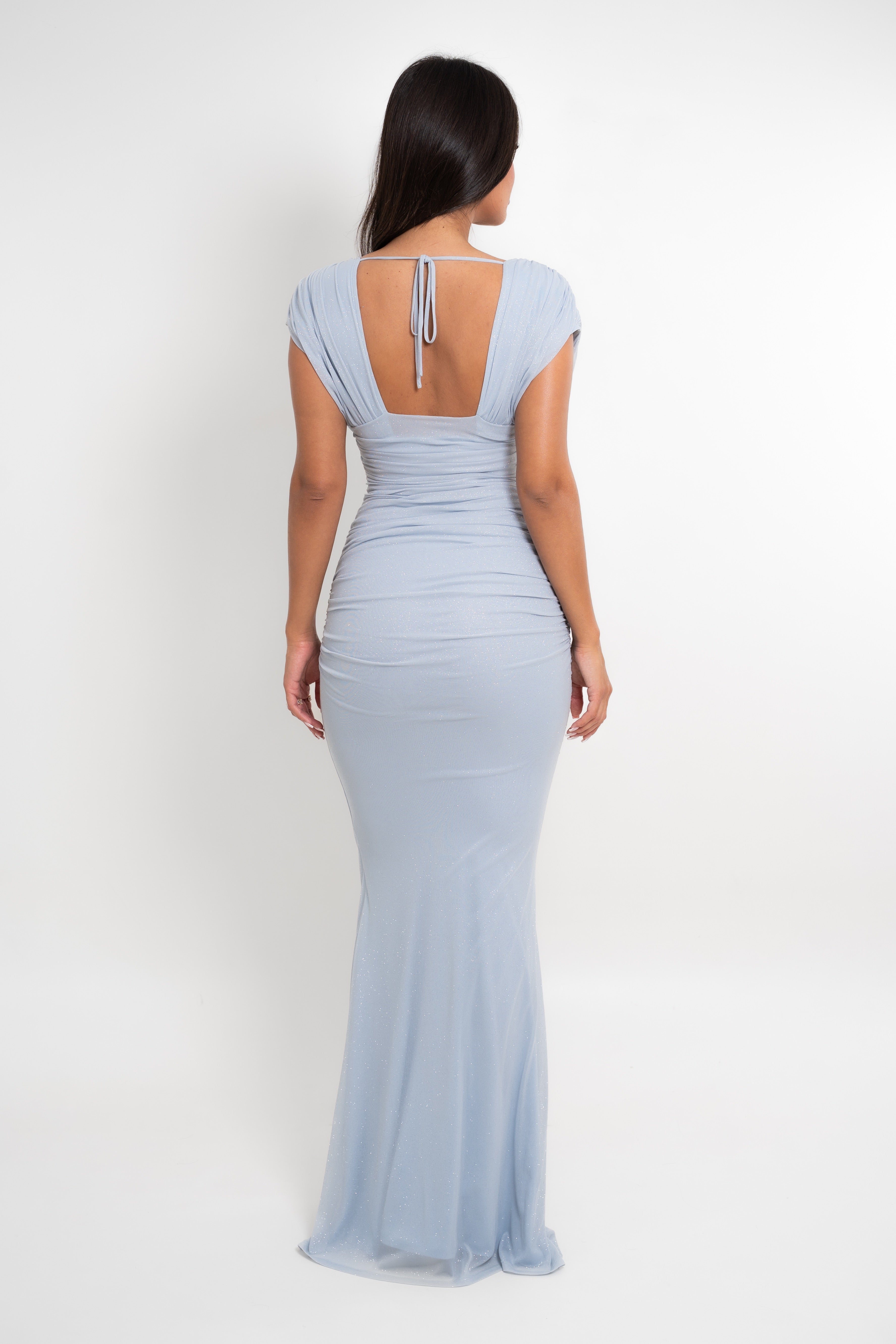 A woman viewed from the back wearing a light blue floor-length gown with cap sleeves, a draped ruched bodice, and a tie detail at the back neckline, against a plain light gray studio background.