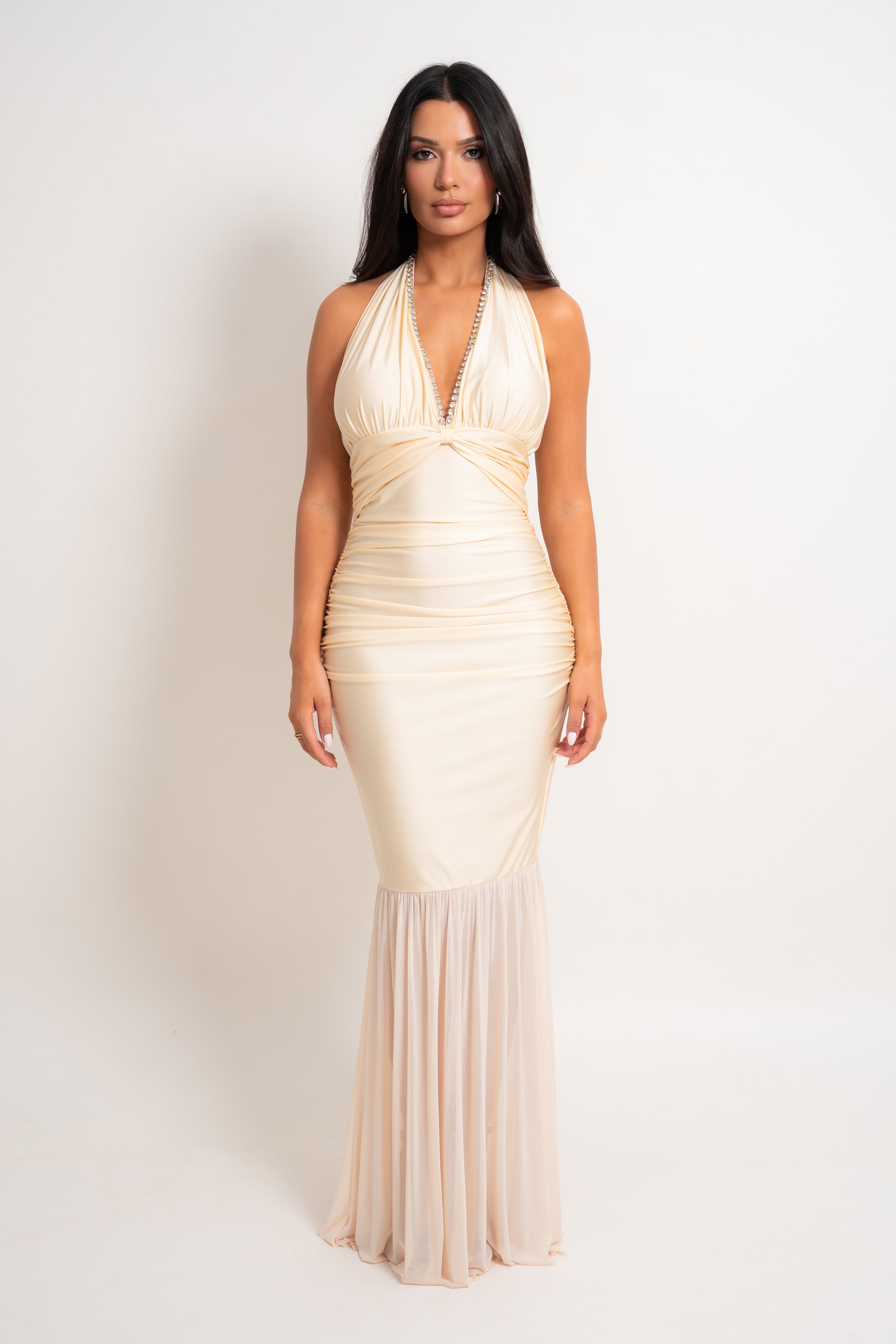 A woman modeling a champagne-colored halter-neck mermaid gown with ruched detailing and a sheer mesh skirt, set against a plain white studio background.