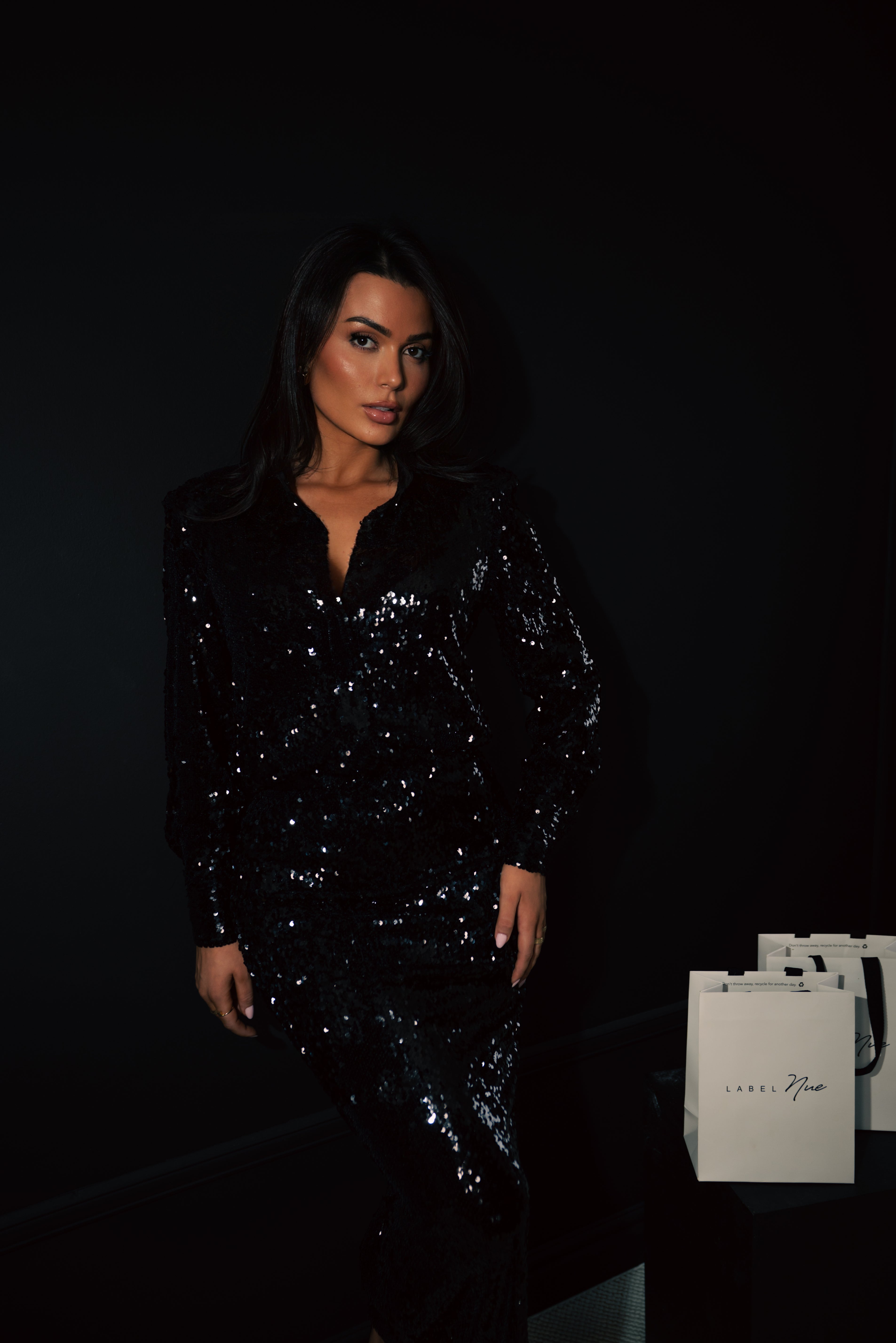 Woman in a black sequin dress standing next to white bags with visible branding.