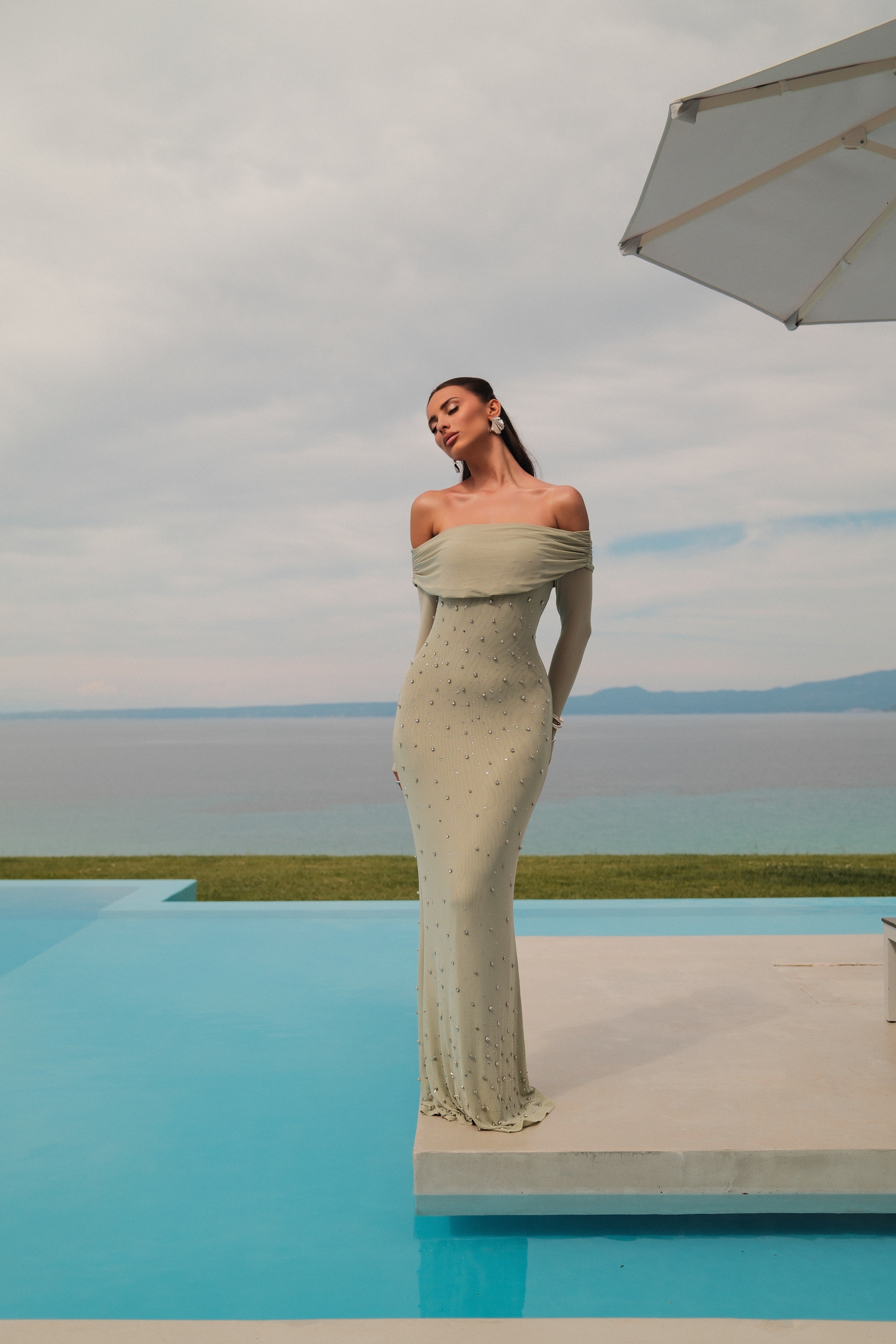 A woman stands on a concrete platform beside a bright blue infinity pool, wearing an off-the-shoulder, floor-length fitted dress with small metallic-looking polka dots. The background shows an outdoor coastal setting with water, distant land, and a cloudy sky, with part of a patio umbrella visible in the upper right.