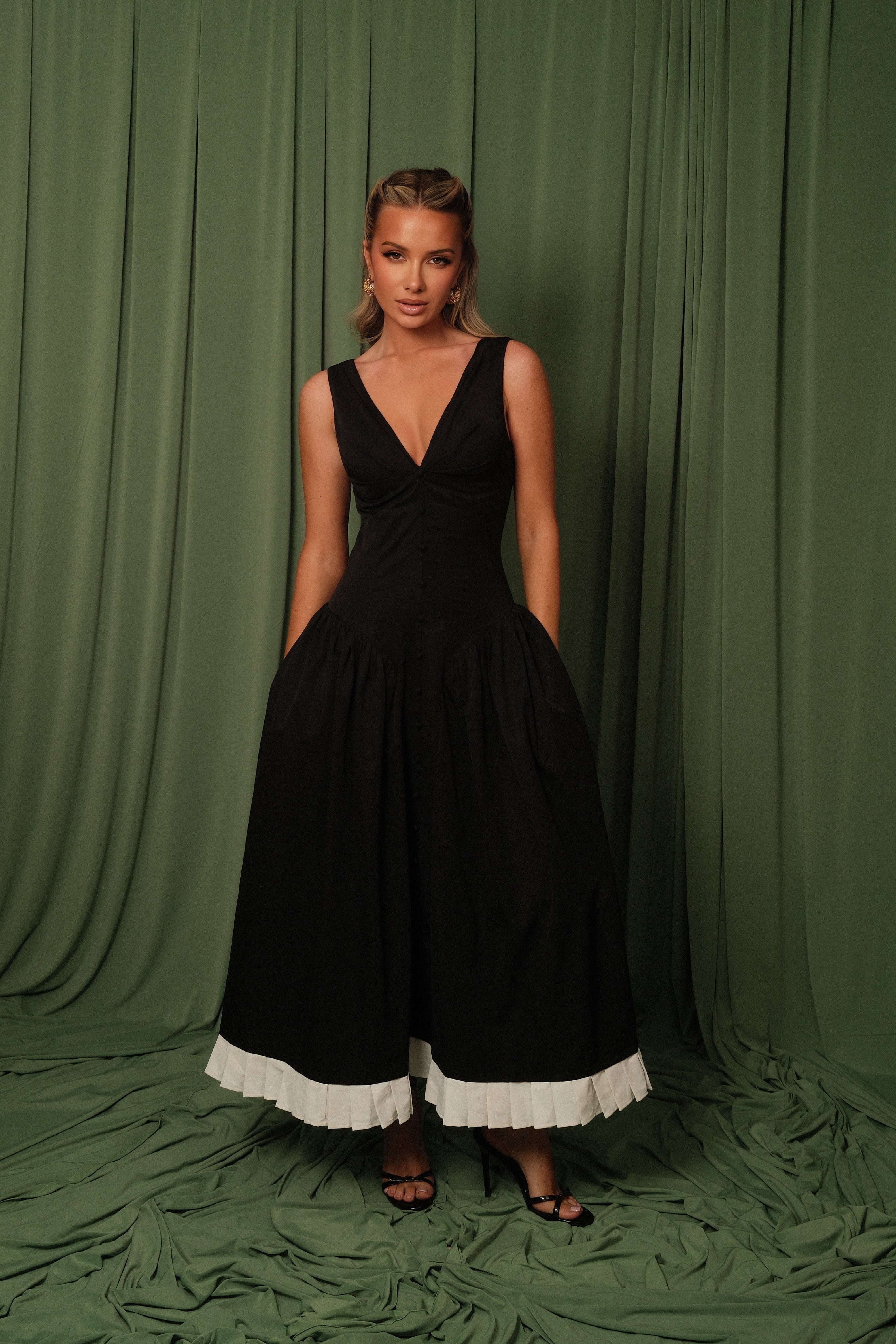 A woman modeling a sleeveless black midi dress with a deep V-neckline, gathered skirt, and a contrasting white ruffled hem, posed against a draped olive green fabric backdrop.