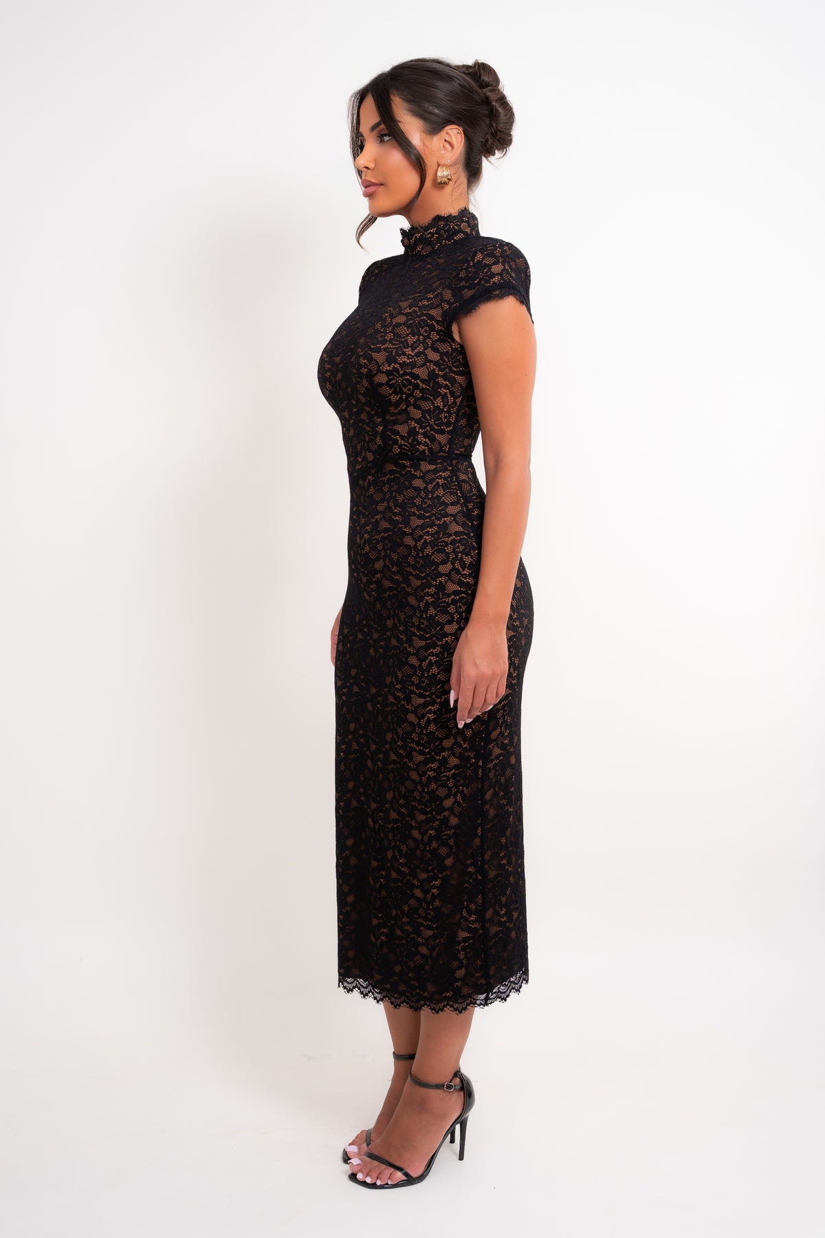 A woman modeling a black lace midi dress with a mock neck and cap sleeves, standing in profile against a plain white studio background.