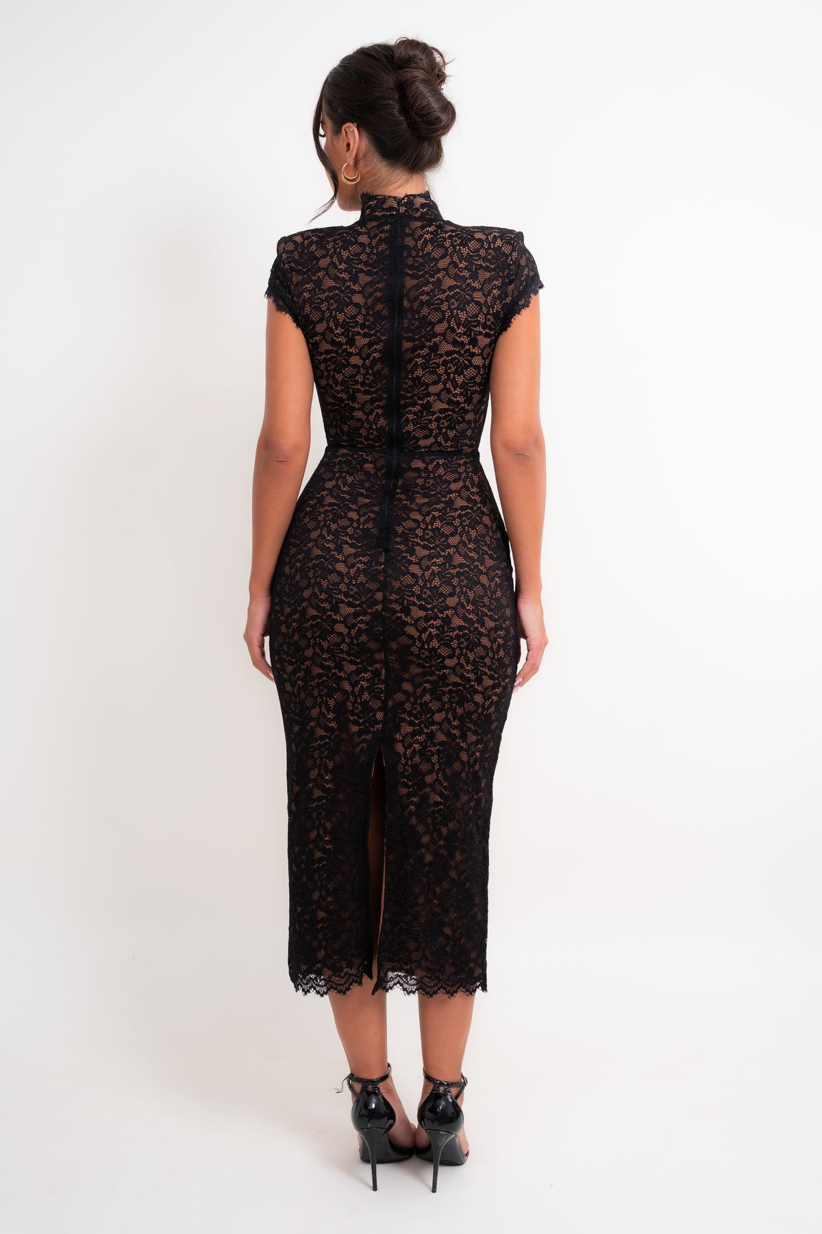 A woman viewed from the back wearing a black lace midi dress with cap sleeves, a high mock neck, and a center back zipper, paired with black high heels against a plain white studio background.