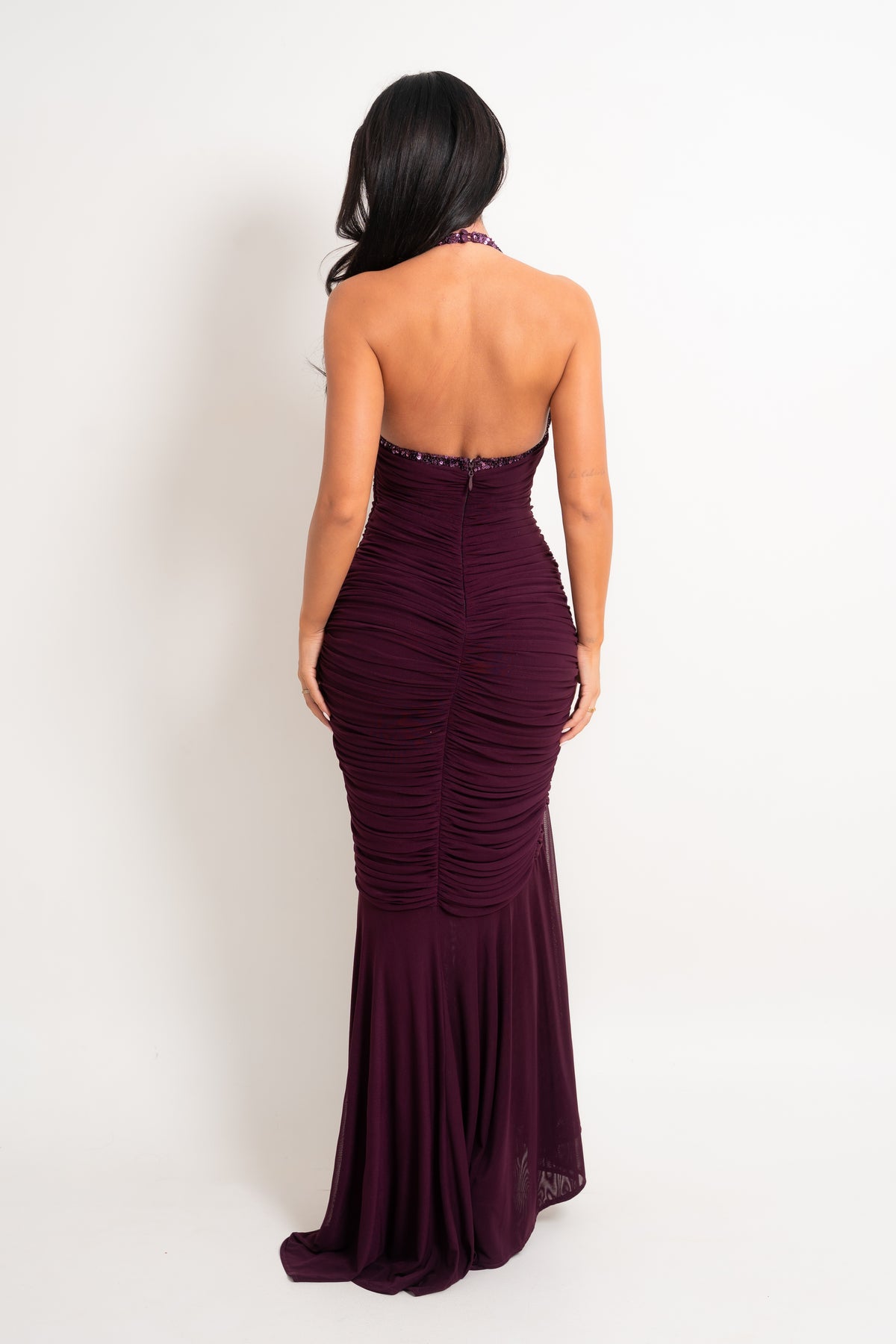 A woman wearing a deep burgundy backless ruched mermaid gown, photographed from behind against a plain white studio background.