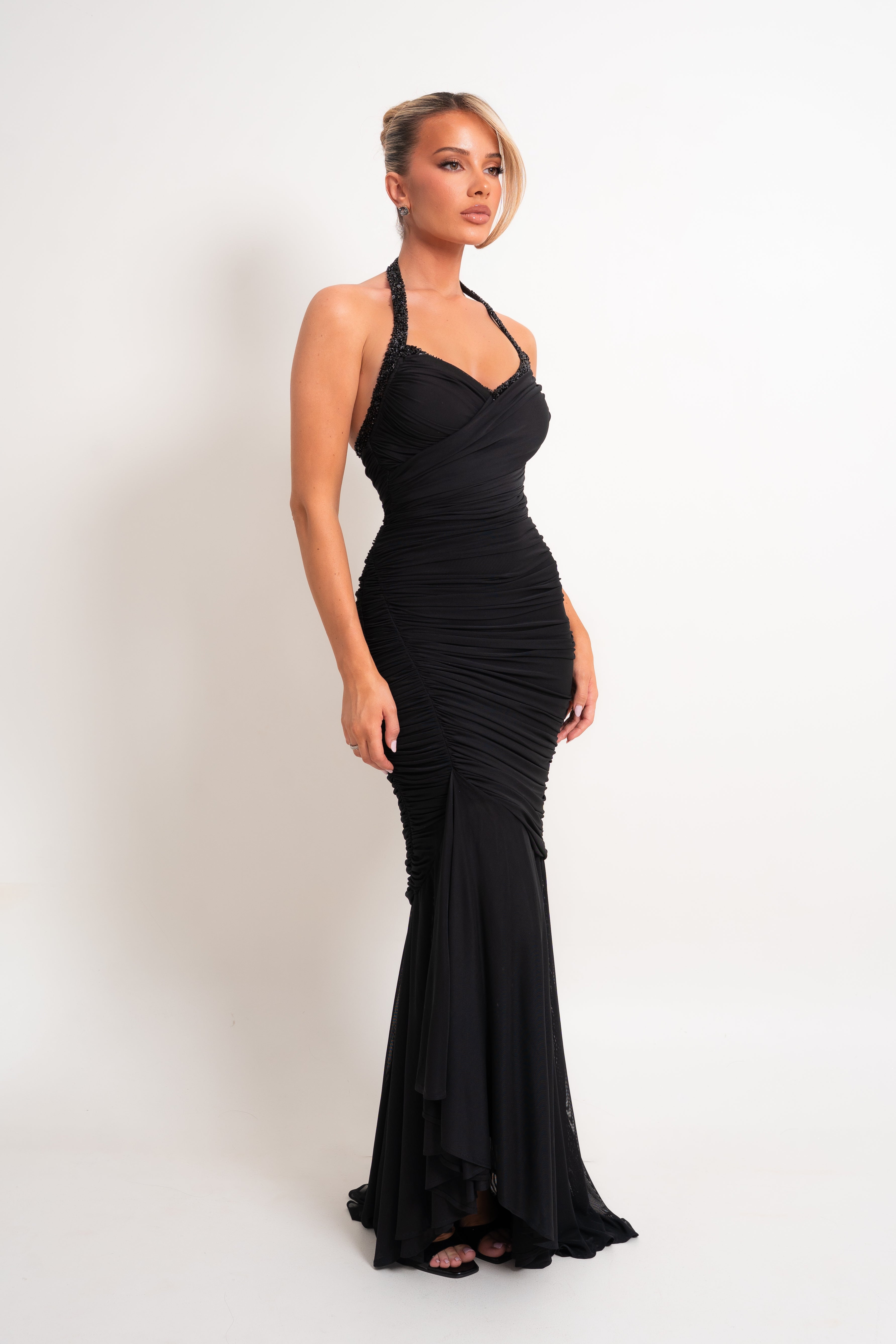A woman modeling a black halter-neck, ruched mermaid gown against a plain white studio background.