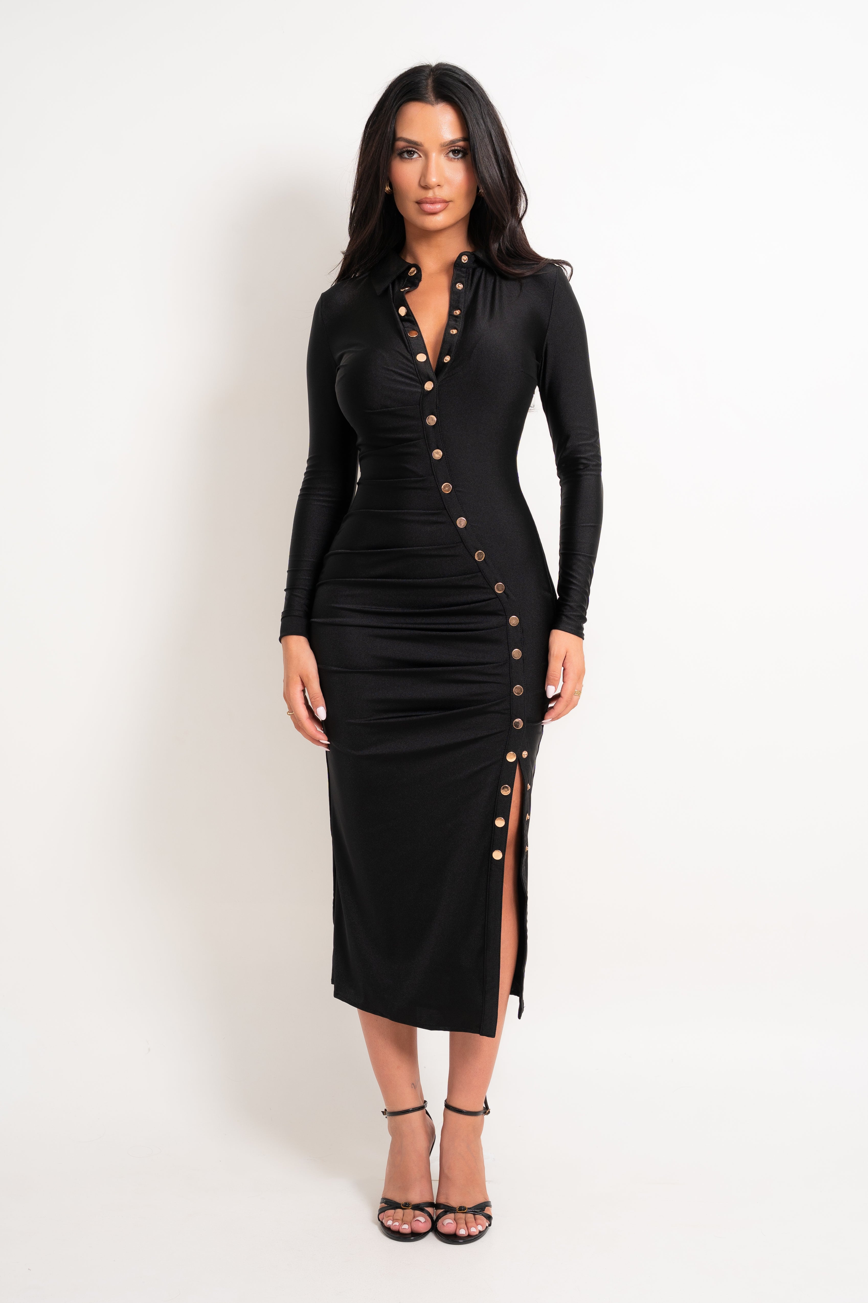 A woman modeling a fitted black long-sleeve midi dress with gold-tone decorative buttons down the front and side slit, photographed against a plain light background.