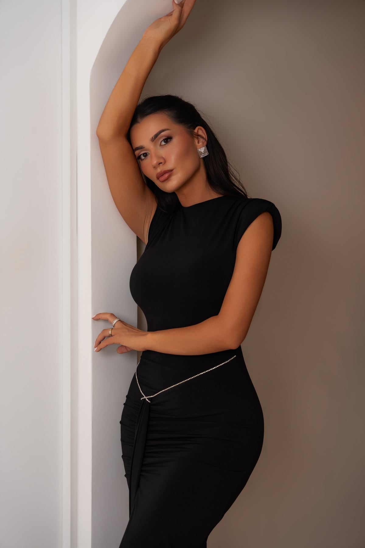 A woman modeling a sleeveless black bodycon dress with a draped wrap detail at the waist, posed against a plain beige background.