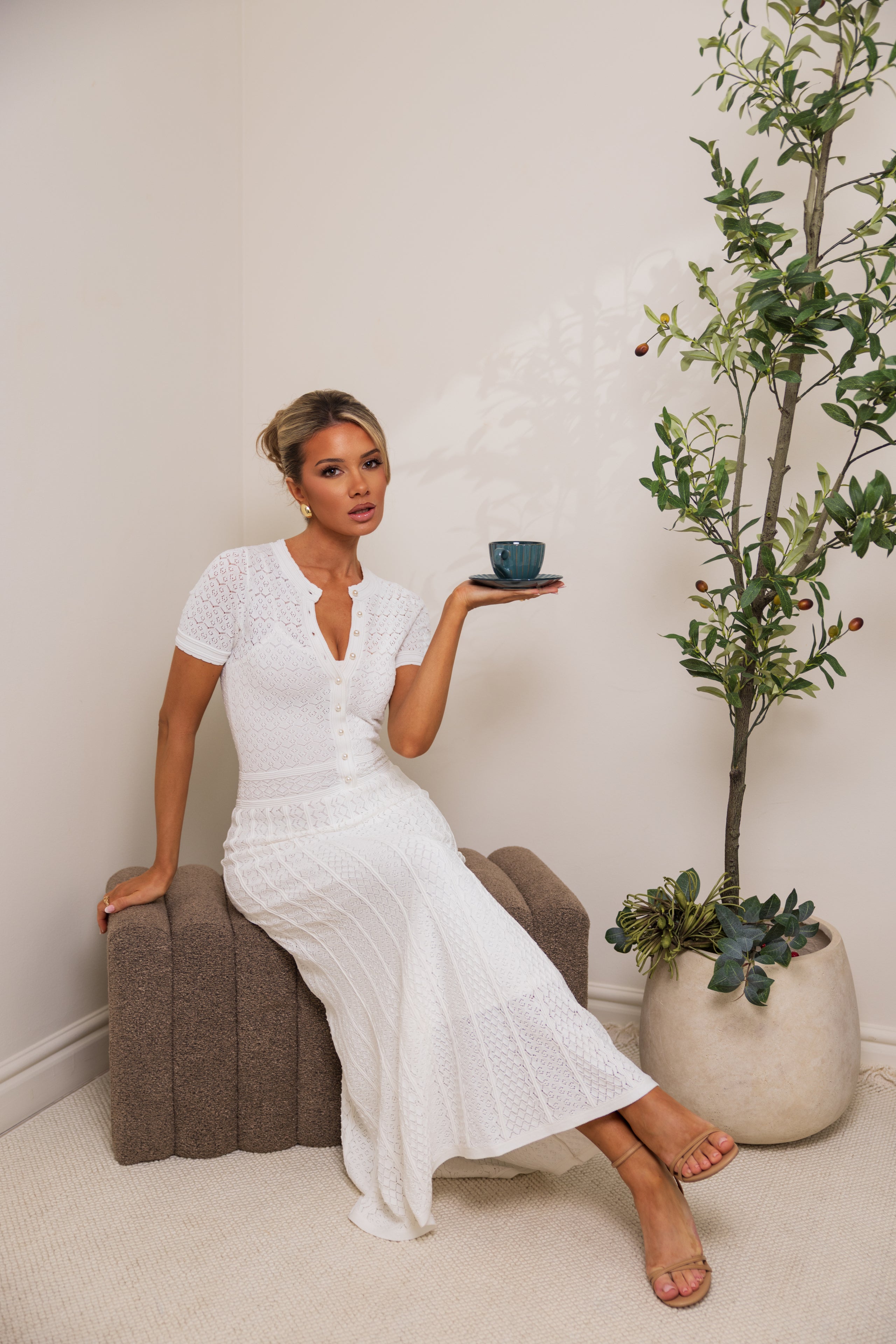 Woman in a white dress holding a small bowl next to a potted plant indoors.
