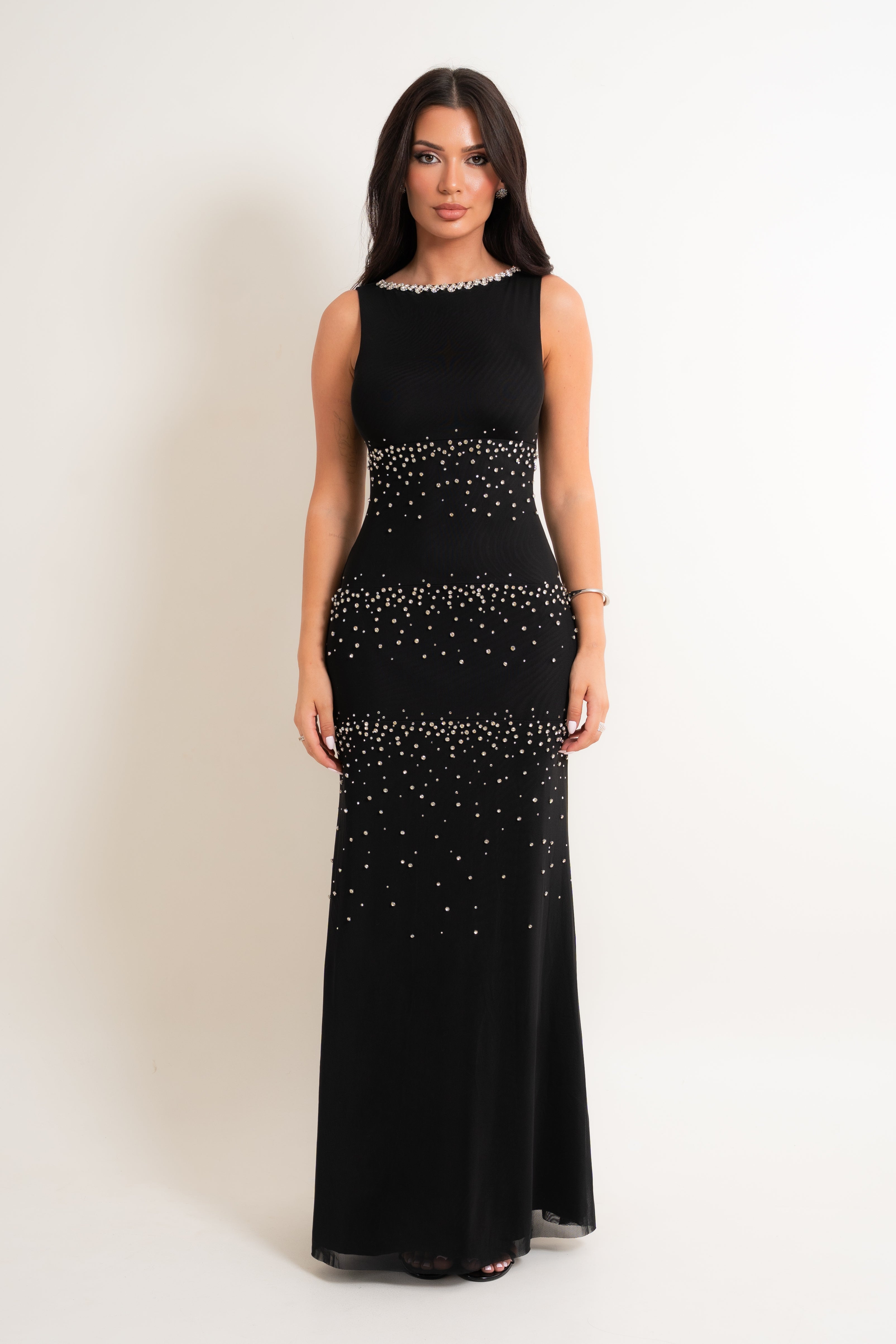 A woman modeling a sleeveless black floor-length gown with beaded horizontal embellishment details across the bodice, waist, hips, and skirt, photographed against a plain light background.