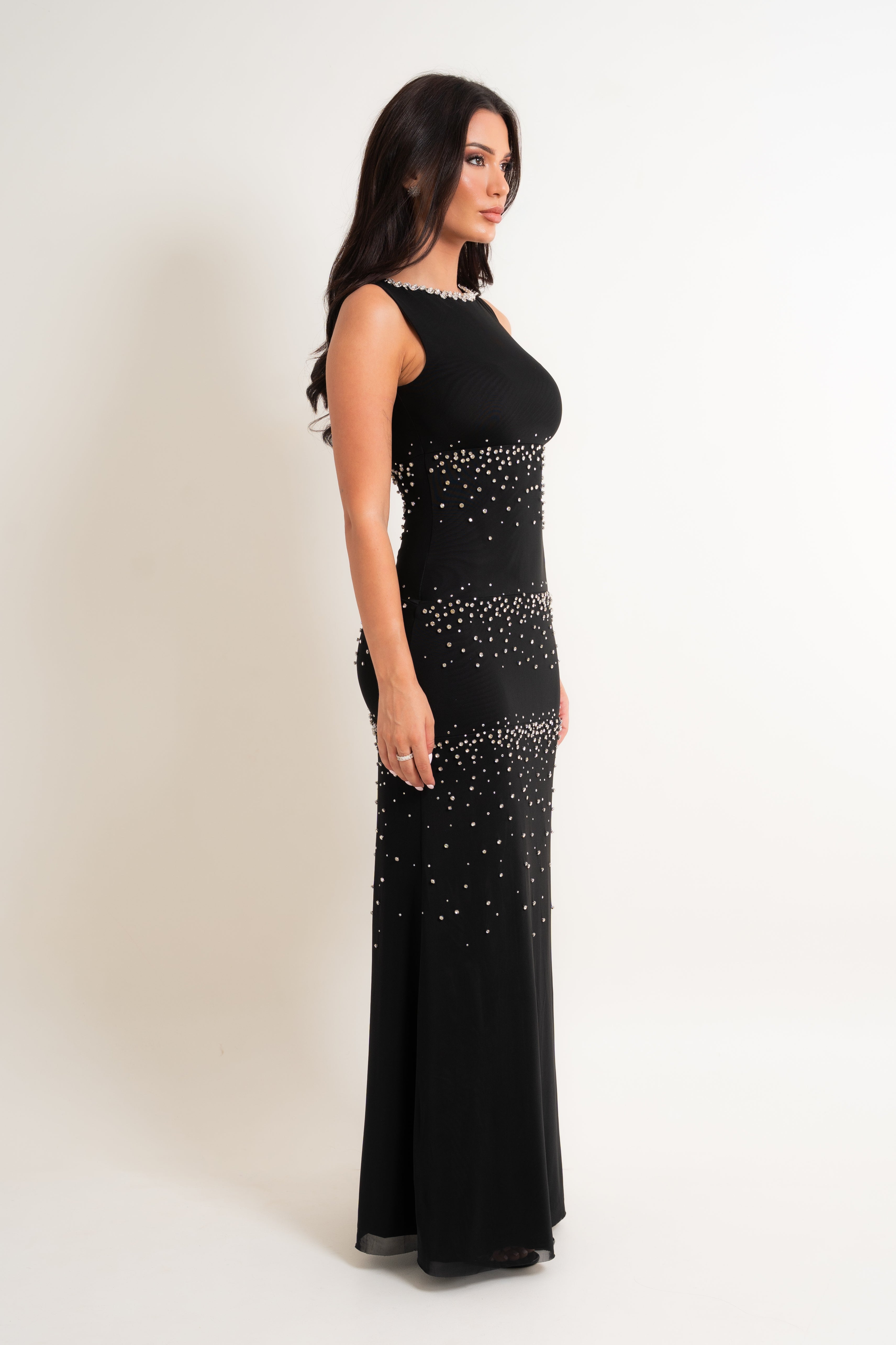 A woman modeling a sleeveless black floor-length gown with beaded embellishments, shown in profile against a plain light beige studio background.
