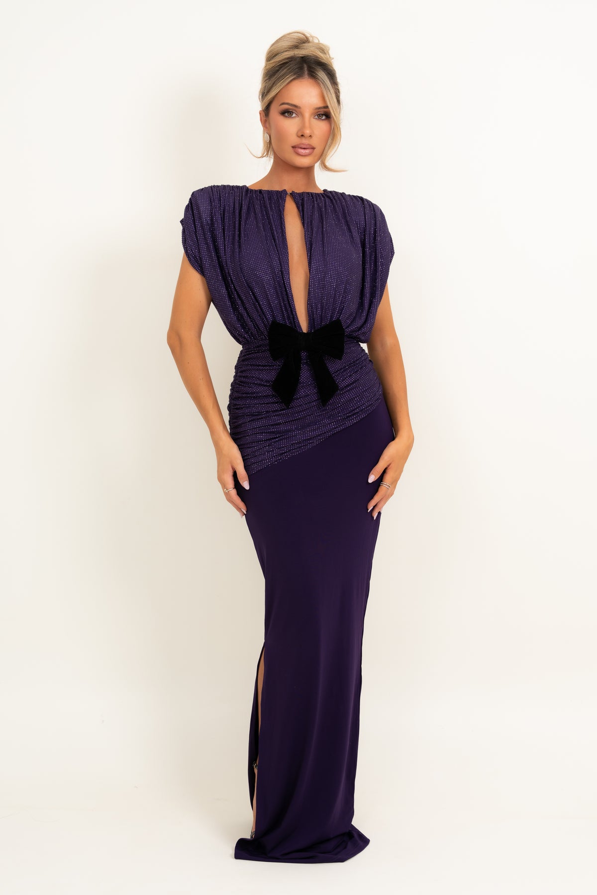 A woman modeling a floor-length, deep purple gown with a plunging keyhole neckline, gathered ruching, a black bow at the waist, and a thigh-high slit, set against a plain white studio background.