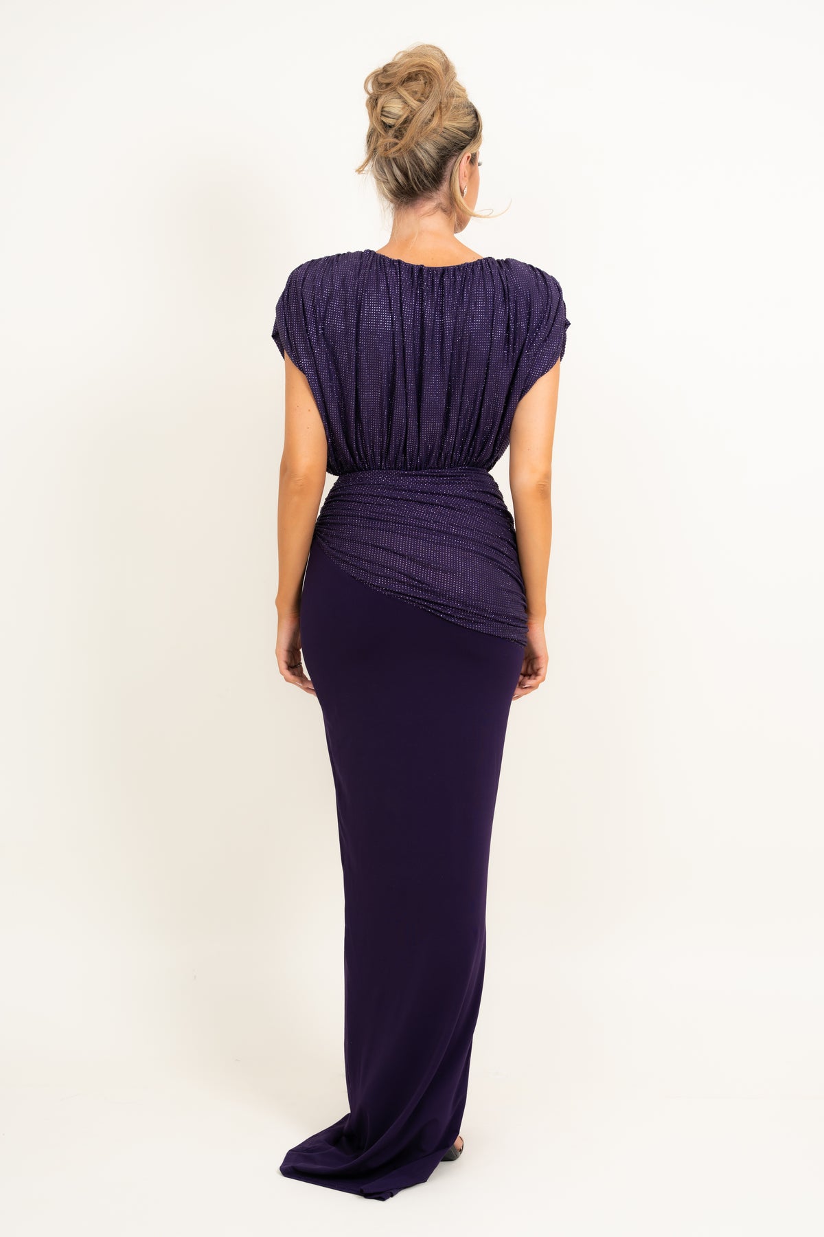 A woman viewed from the back wearing a floor-length, deep purple gown with short cap sleeves, a gathered bodice, and draped detailing around the waist, photographed against a plain light background.