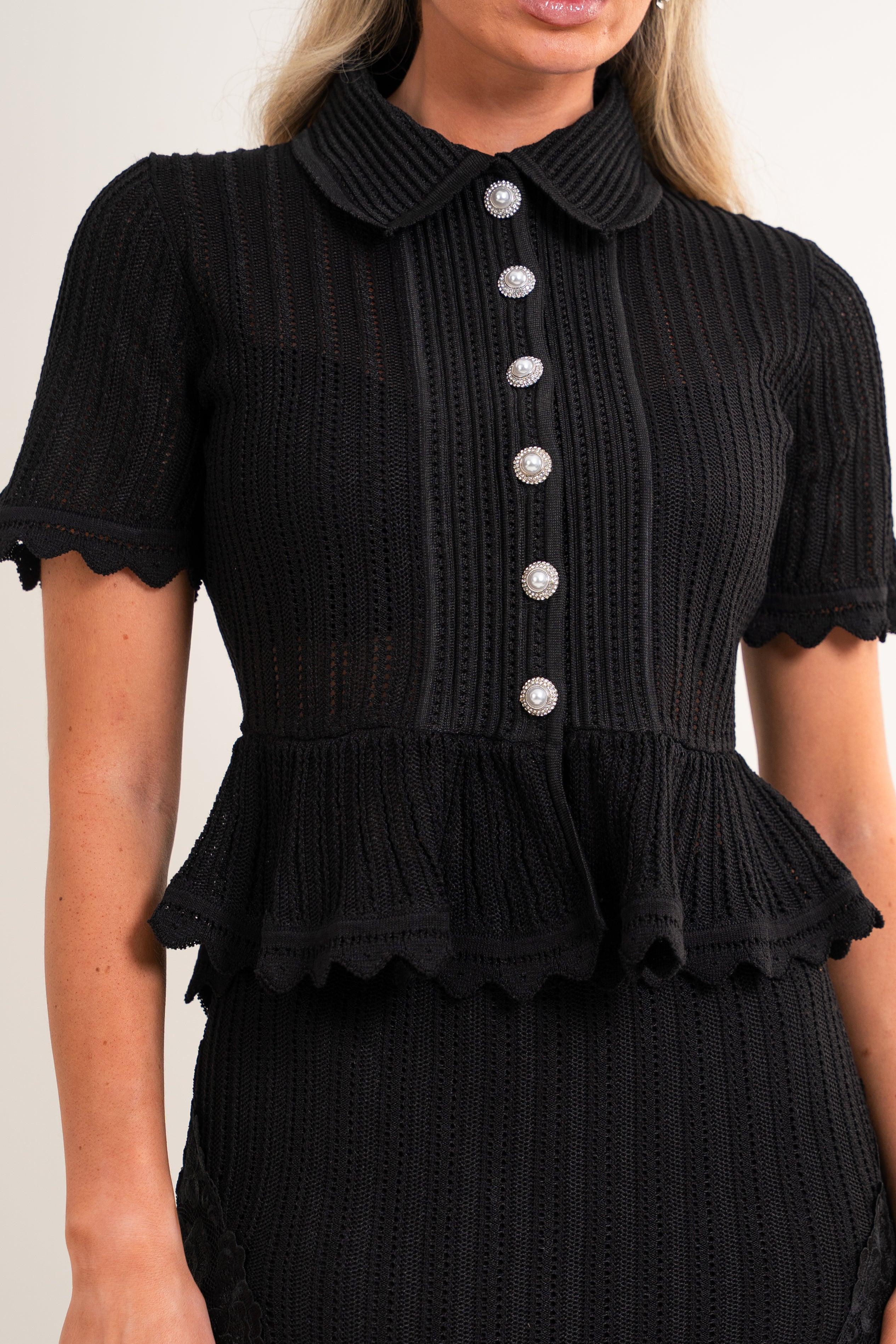 A close-up of a woman wearing a black short-sleeve ribbed knit top with a Peter Pan collar, scalloped edges, and a vertical row of silver buttons down the front.