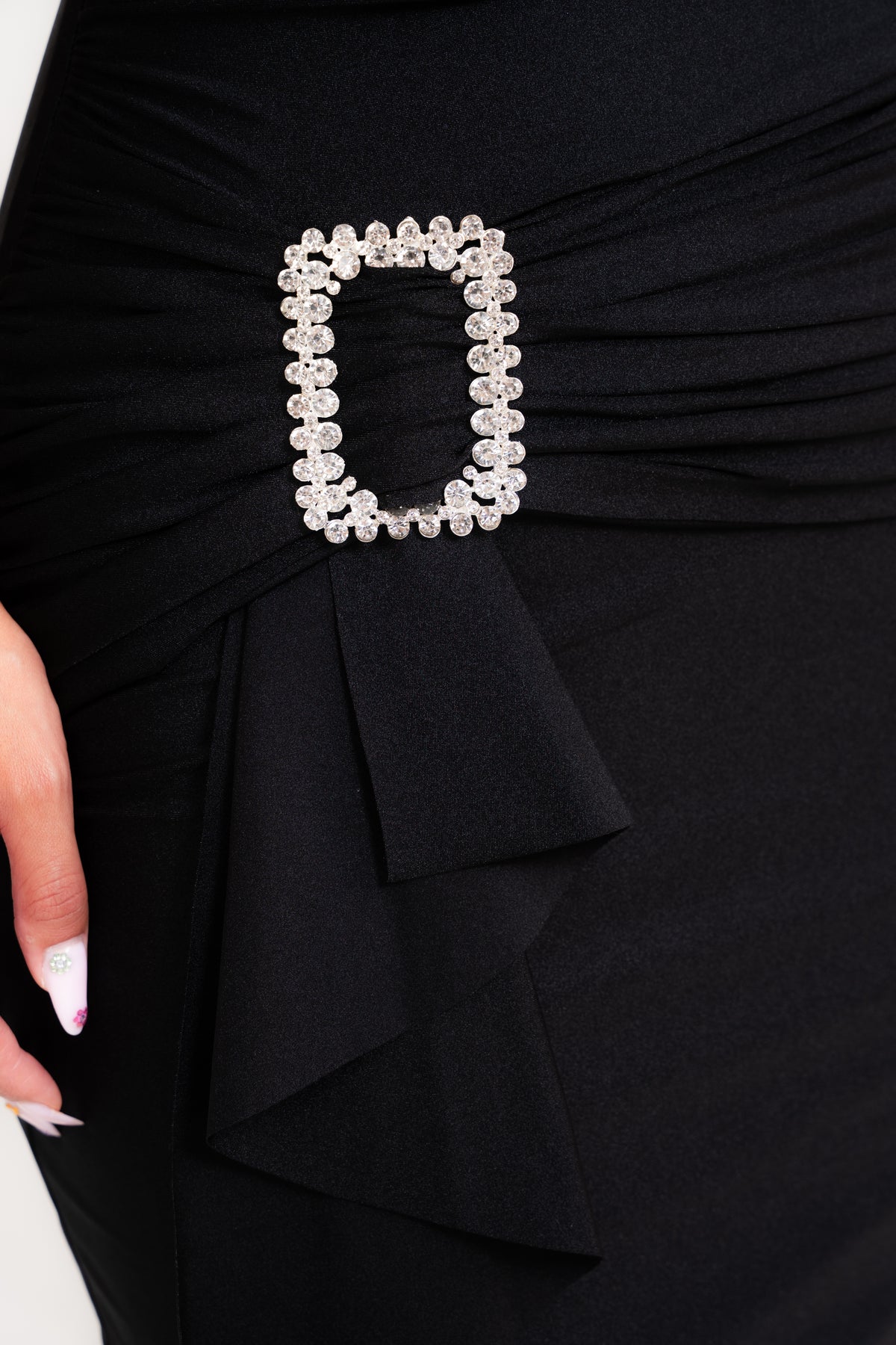 Close-up of a black ruched dress with a decorative rectangular silver rhinestone belt buckle at the waist, with a hand partially visible on the left.