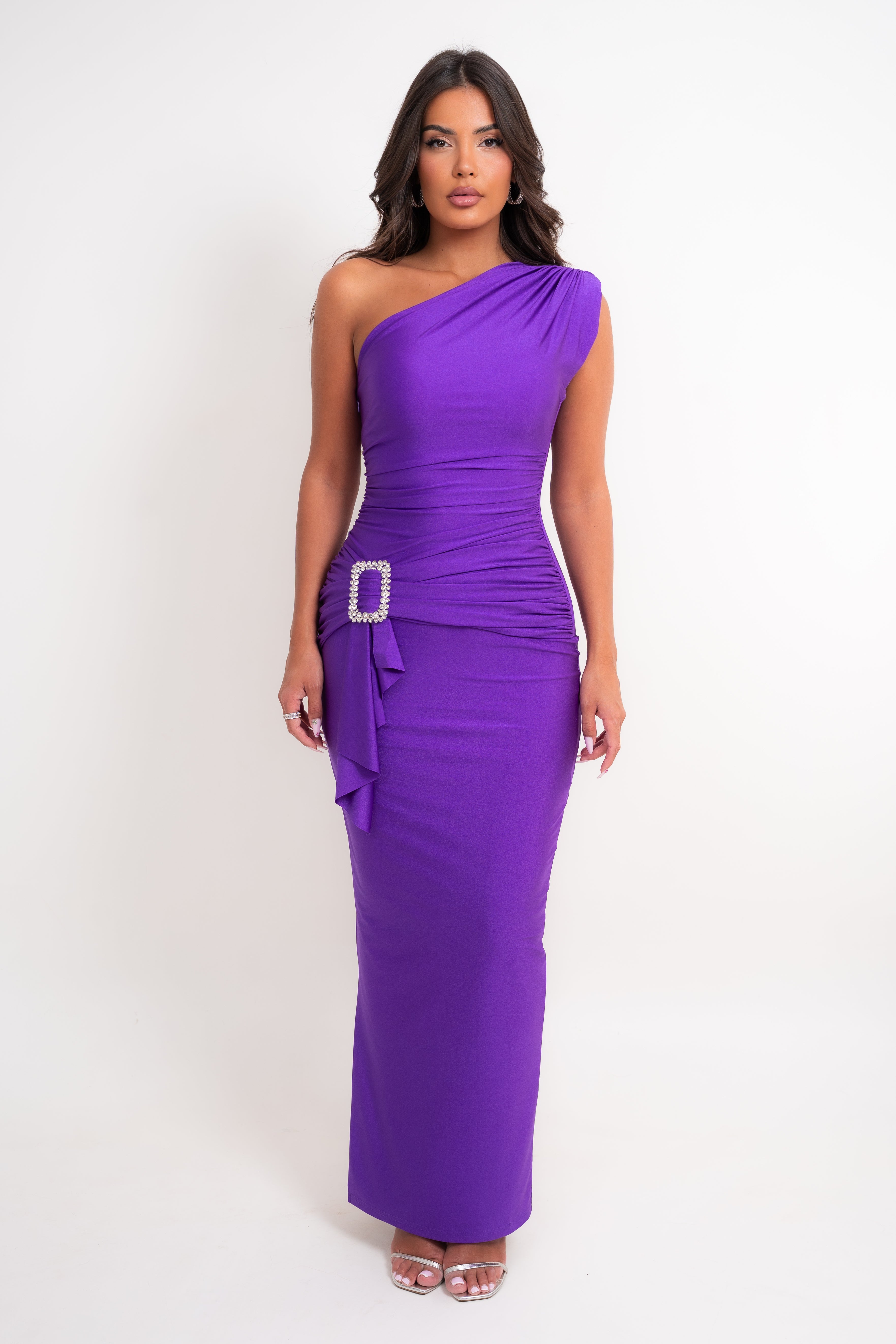 A woman modeling a vibrant purple one-shoulder maxi dress with ruching, a side tie detail, and a rhinestone-embellished belt buckle, set against a plain white studio background.