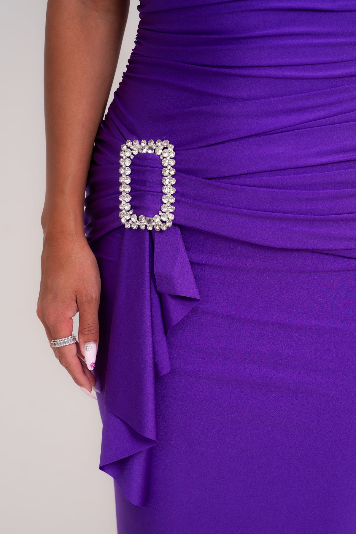 Close-up of a model wearing a ruched purple dress with a draped side detail and a rhinestone-embellished rectangular buckle at the waist.