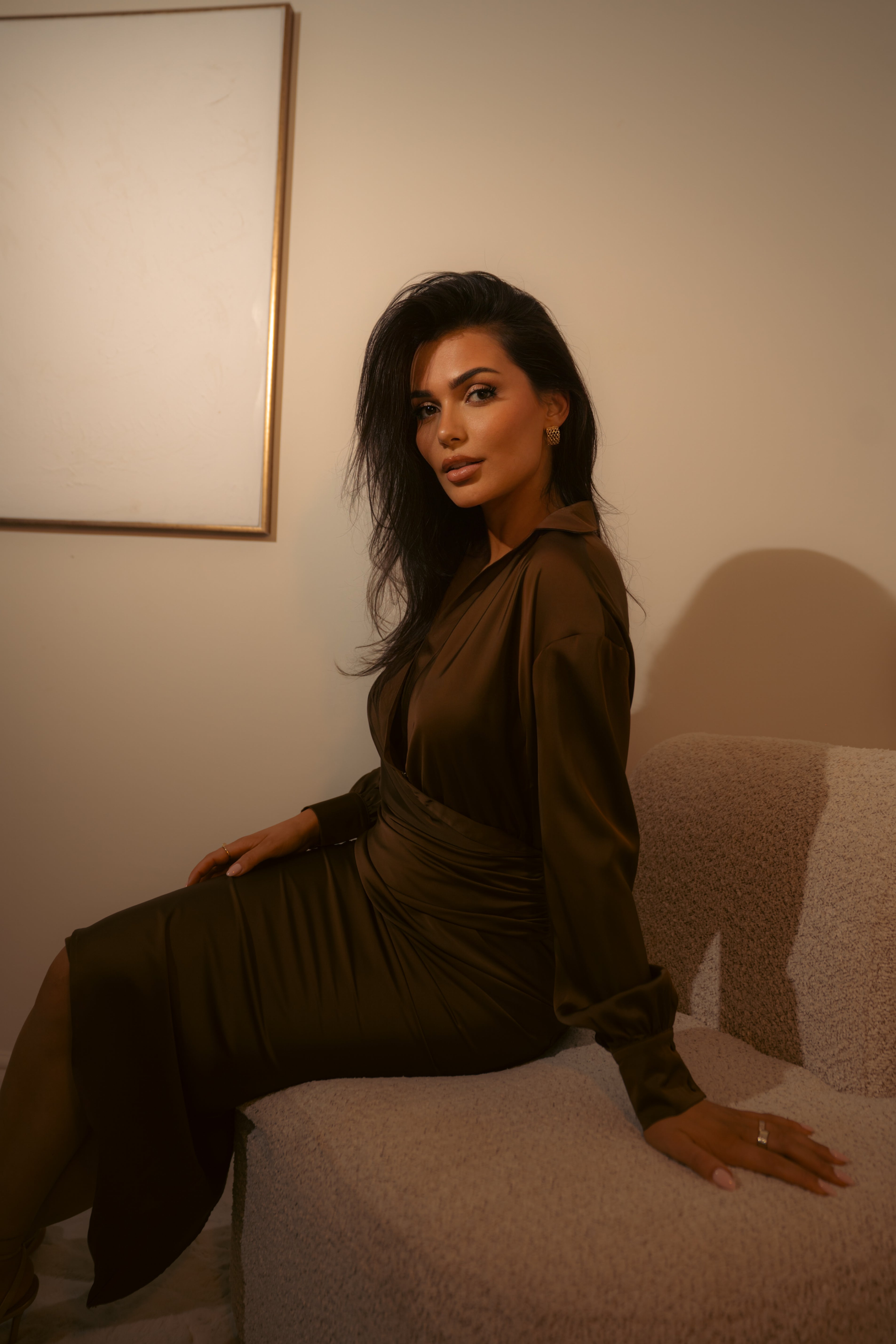 Woman in a brown dress sitting on a couch in a room with a neutral wall.