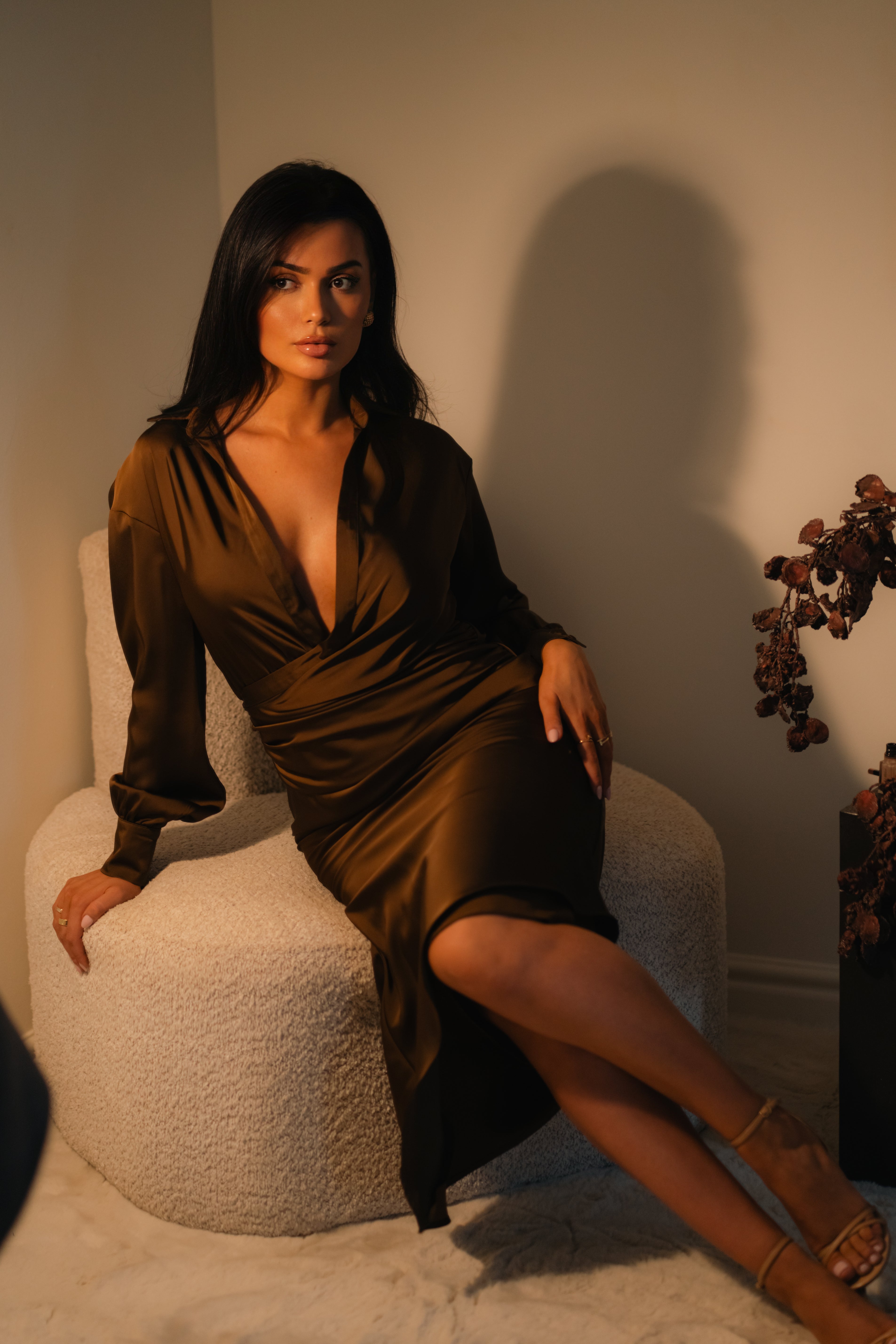 Woman in a brown dress sitting on a white chair in a softly lit room.