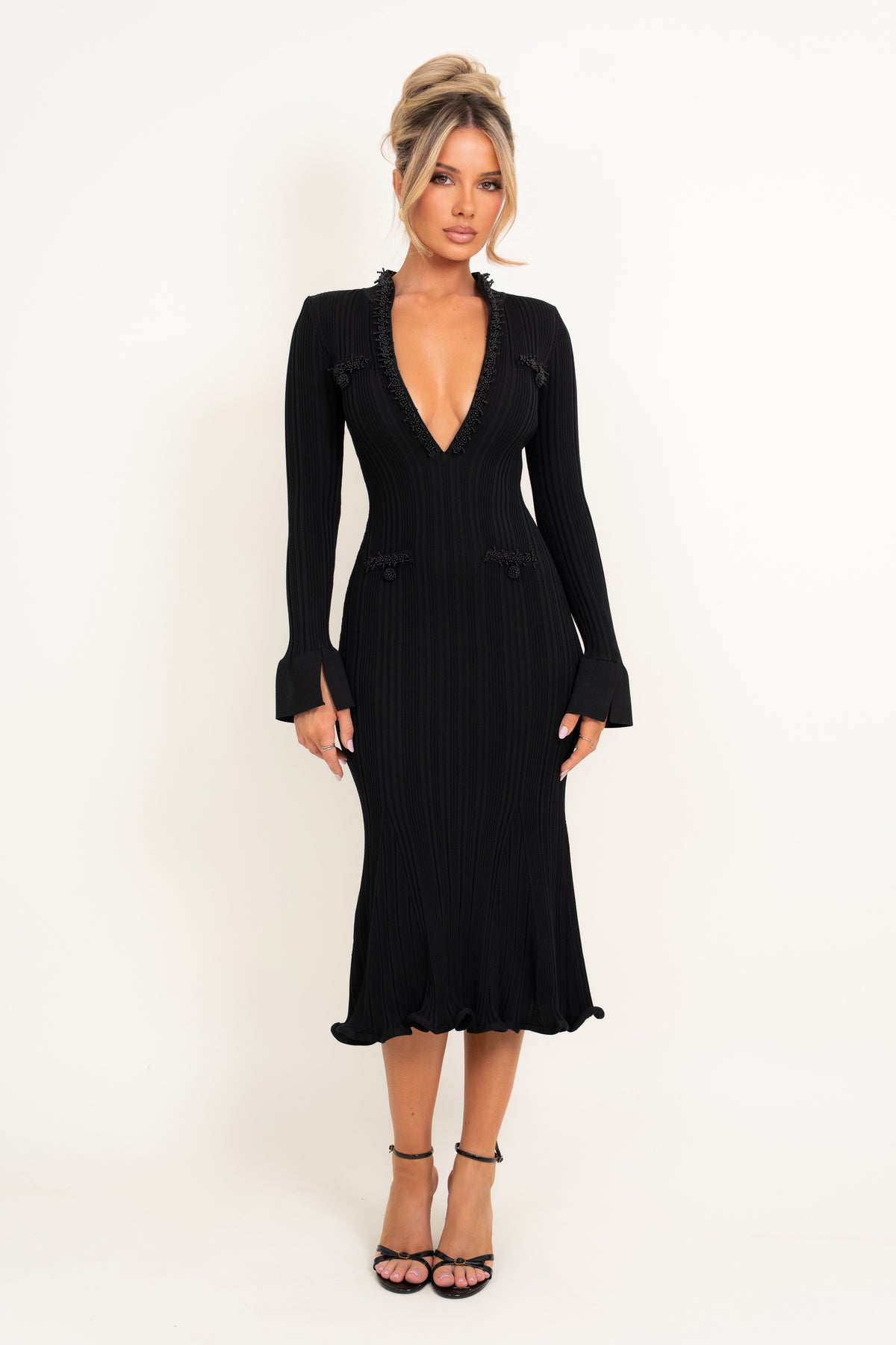 A woman modeling a black long-sleeve ribbed midi dress with a deep V-neckline, ruffled trim details, and flared cuffs, paired with black strappy heels against a plain white studio background.
