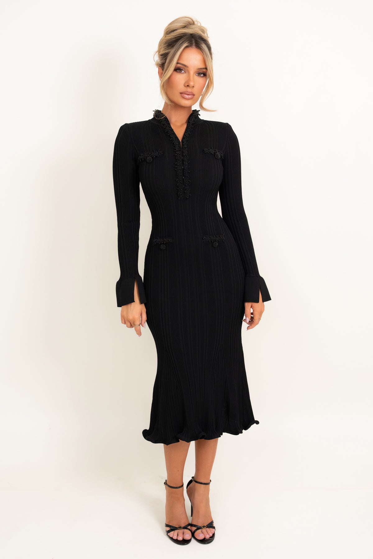 A woman modeling a black long-sleeve midi dress with pleated texture, ruffled trim at the collar and cuffs, and button details on the chest and waist, photographed against a plain white studio background.
