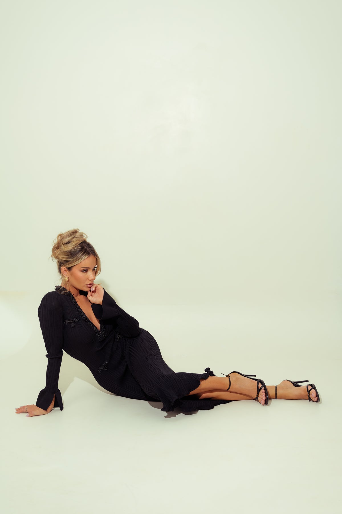 A woman with blonde hair in an updo lies on her side on a white studio floor, wearing a black long-sleeve wrap-style dress with a deep V-neck and black strappy high-heeled sandals.